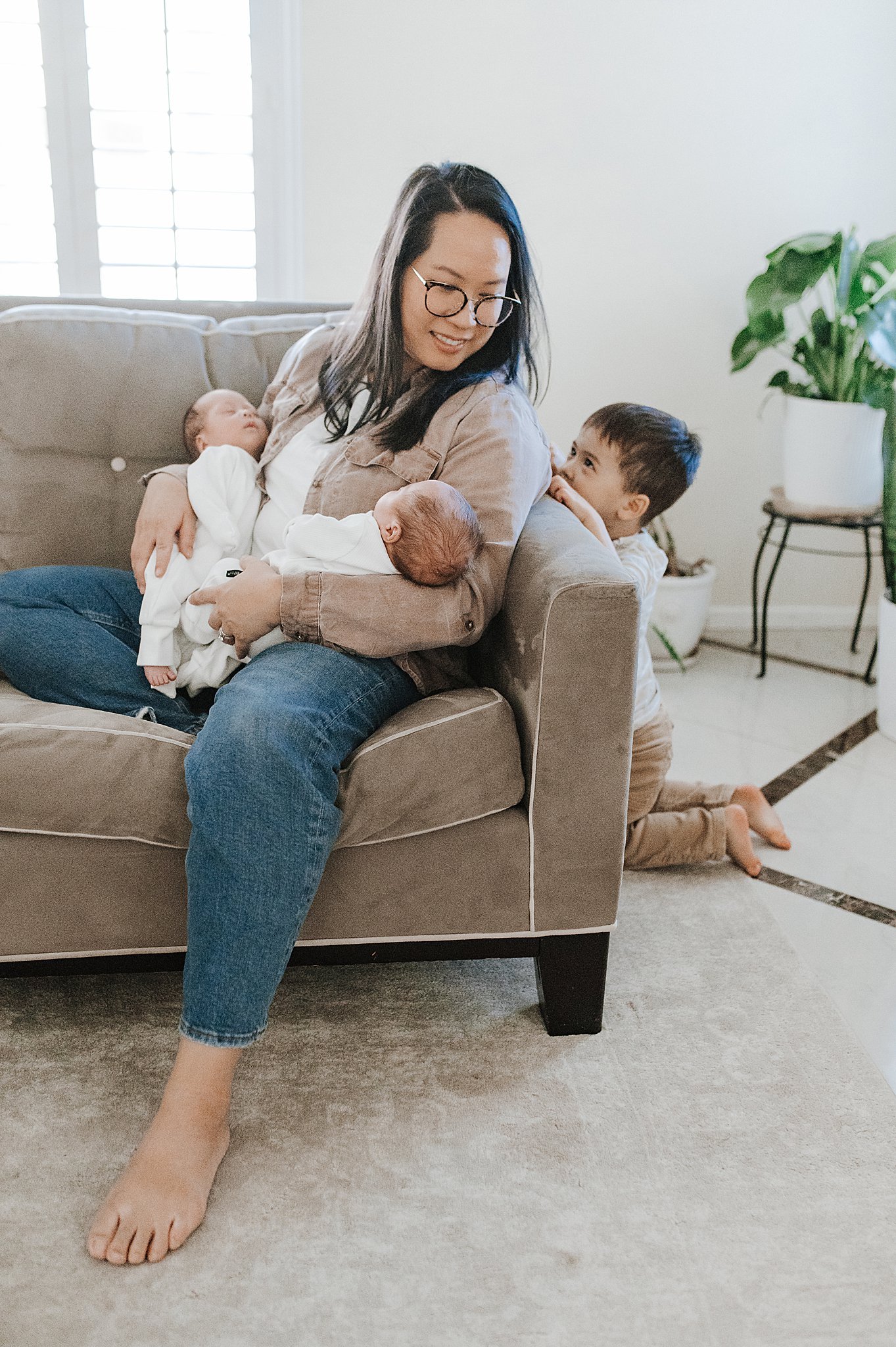 A mom sitting in her living room, holding her two twin baby boys, while her toddler looks on, taken by Alyssa Charboneau Photography.