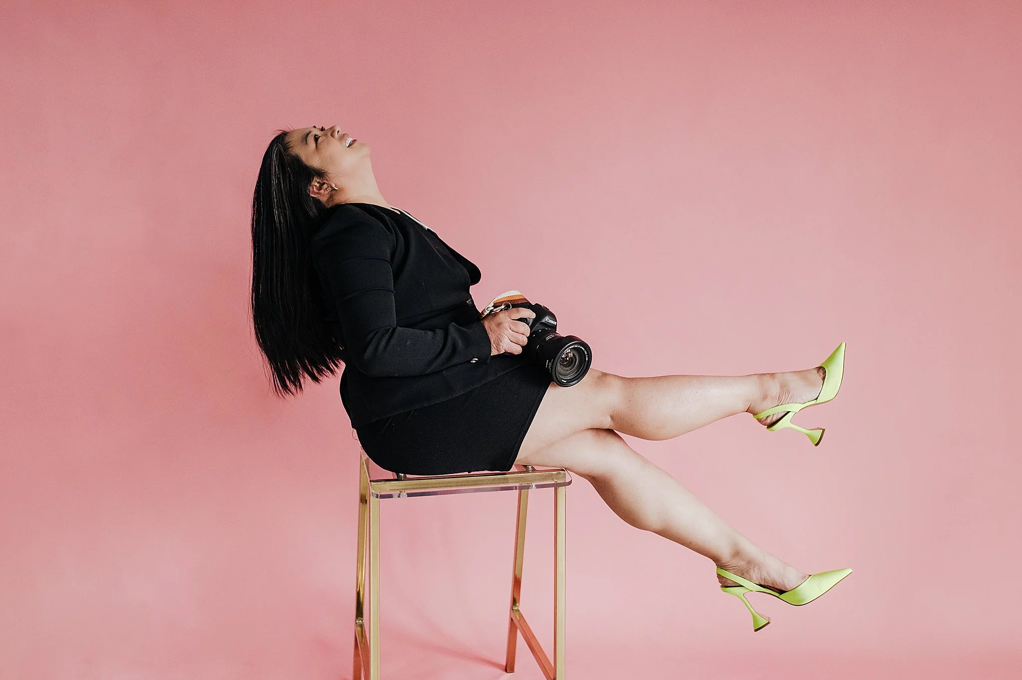 Woman sitting on a transparent chair with a camera in her hand, laughing with her head tilted back, wearing a black blazer, black skirt, and lime green high heels, against a pink background.