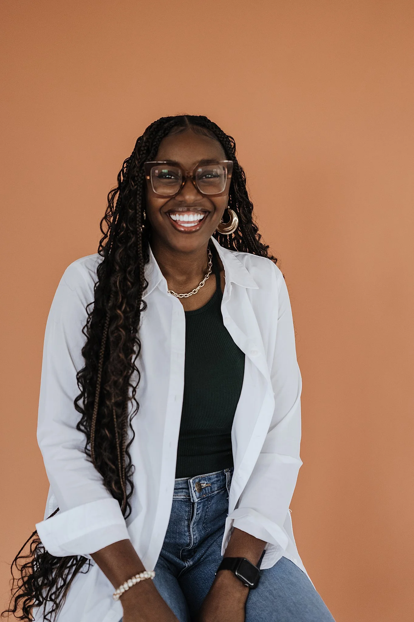 A smiling woman with long curly hair, glasses, and hoop earrings sitting against a peach-colored background. She's wearing a white shirt over a black top, jeans, a black smartwatch, and jewelry including a pearl bracelet, a chain necklace, and earrin