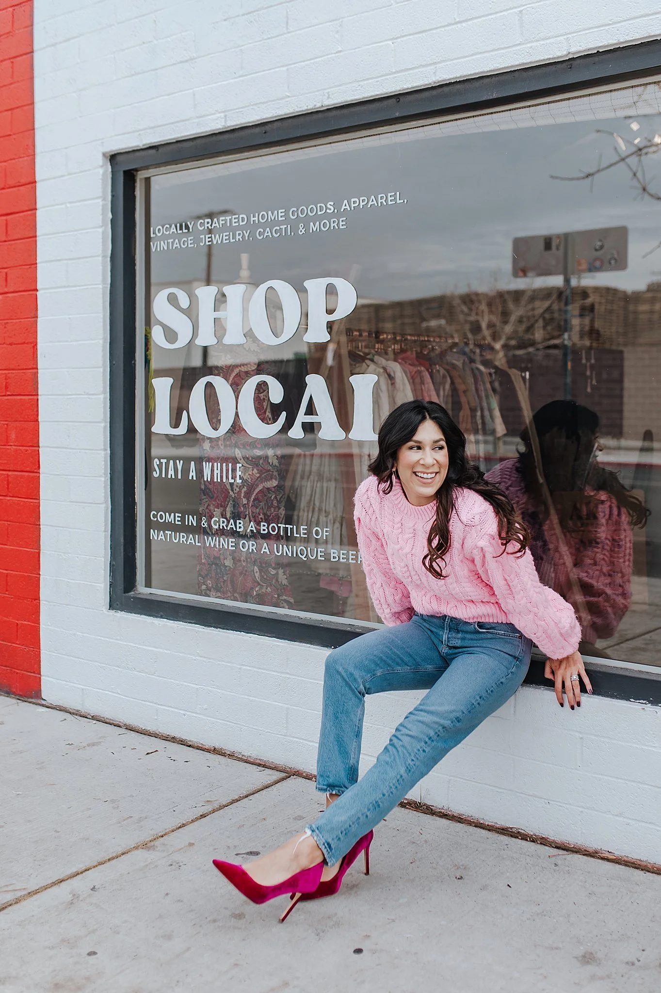 A woman wearing a pink sweater, blue jeans, and hot pink high heels sitting outside a store window that promotes shopping local, with clothing inside the store visible through the glass.