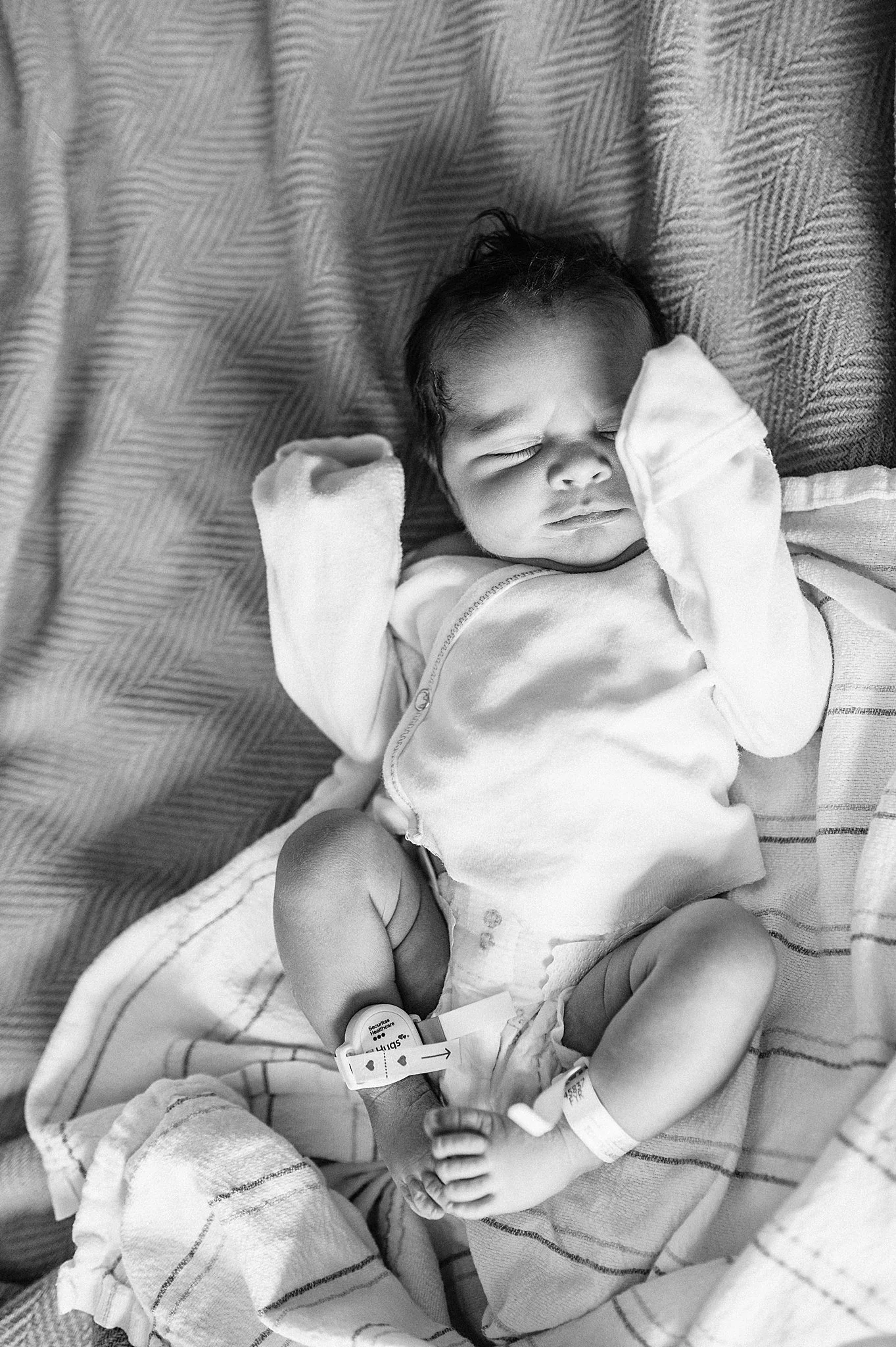 Black and white photo of a sleeping infant lying on a hospital bed wearing a zip-up onesie and blankets covering the body, during a Fresh 48 session at Summerlin Hospital.