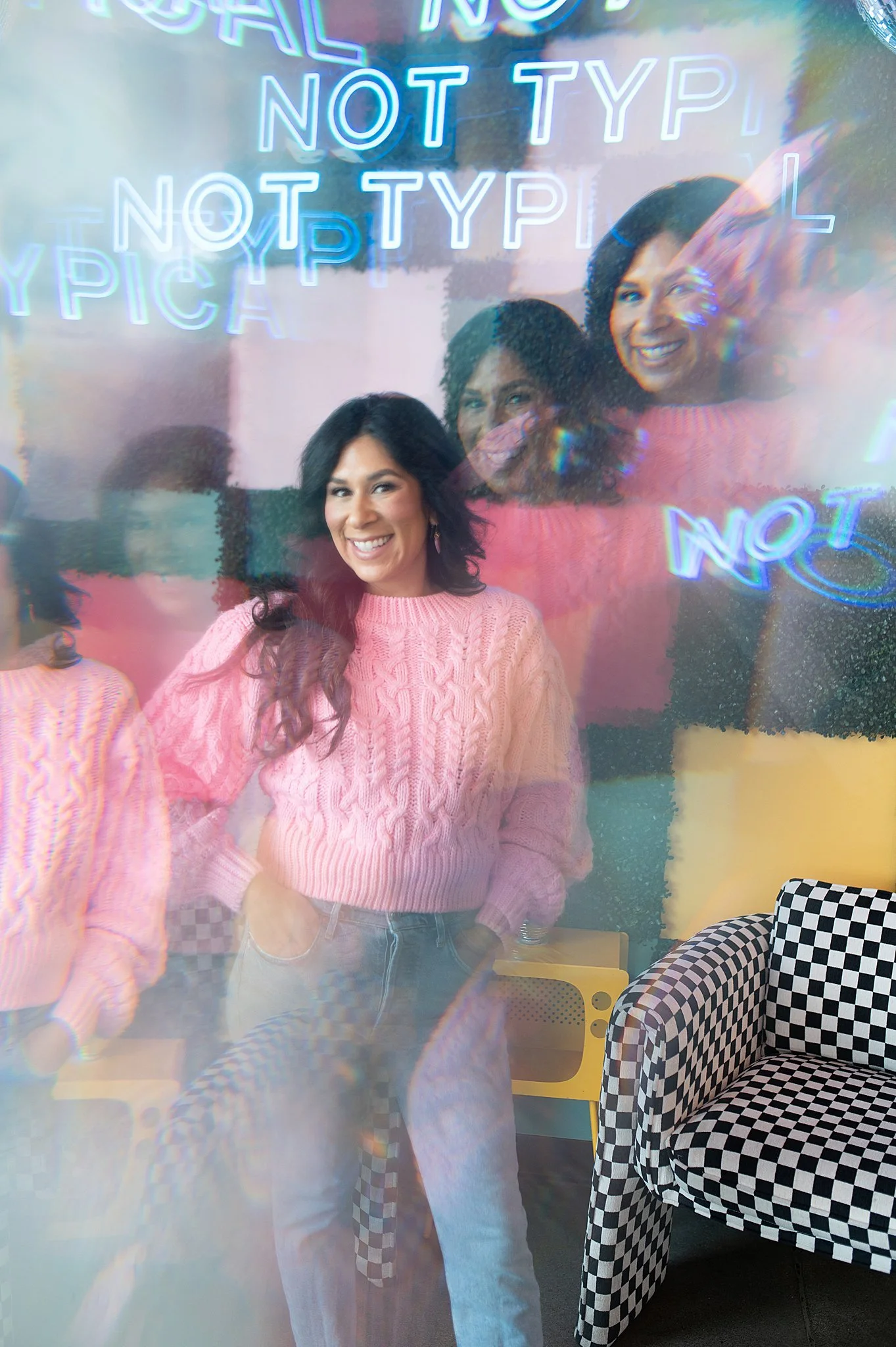 A woman in a pink sweater smiling in front of a colorful wall with neon signs that say 'NOT TYPICAL.' Multiple reflections create a layered effect. Las Vegas branding session.