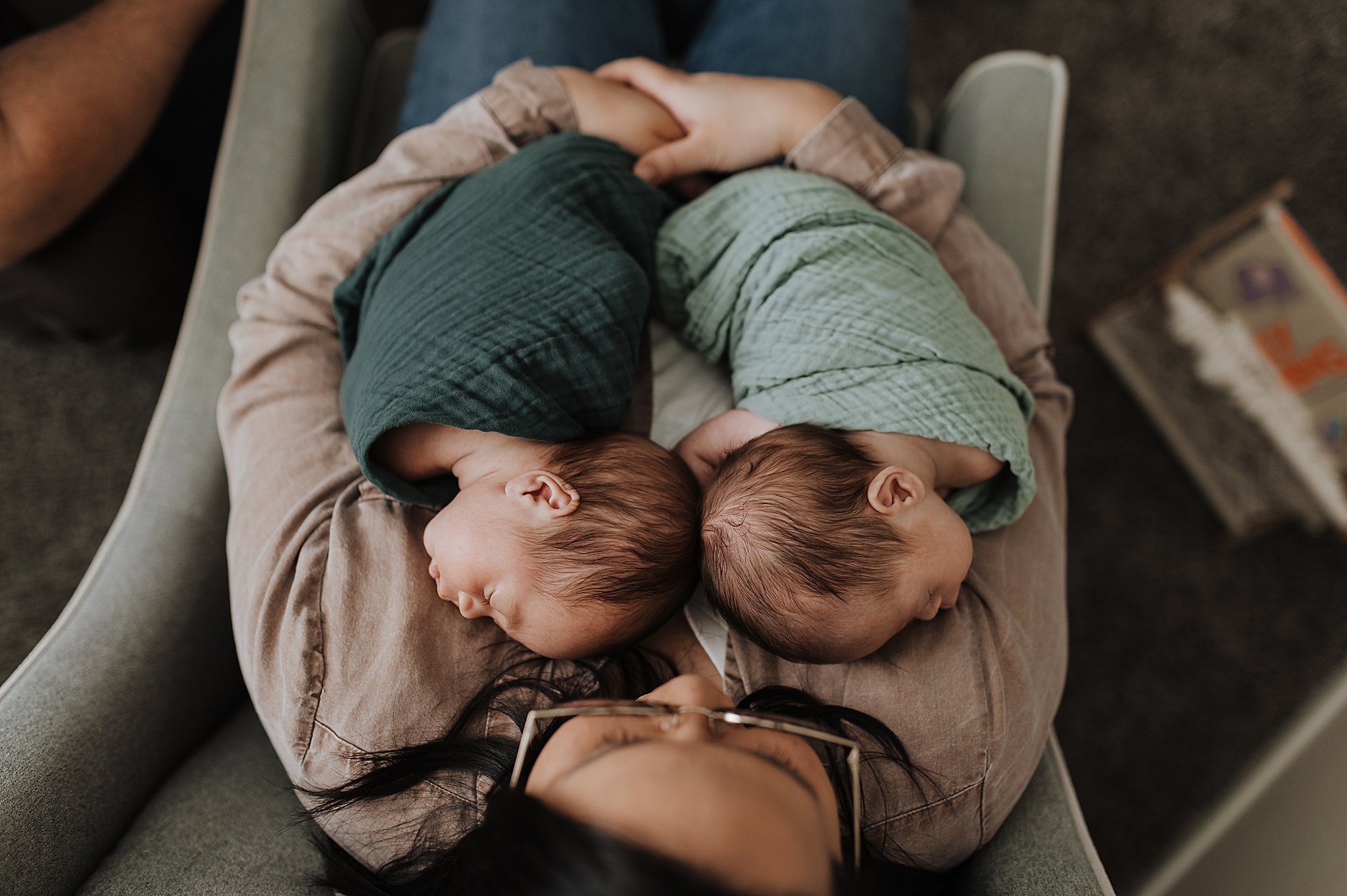 Mother holding newborn twin boys in hunter and sage green swaddles, from an overhead angle.