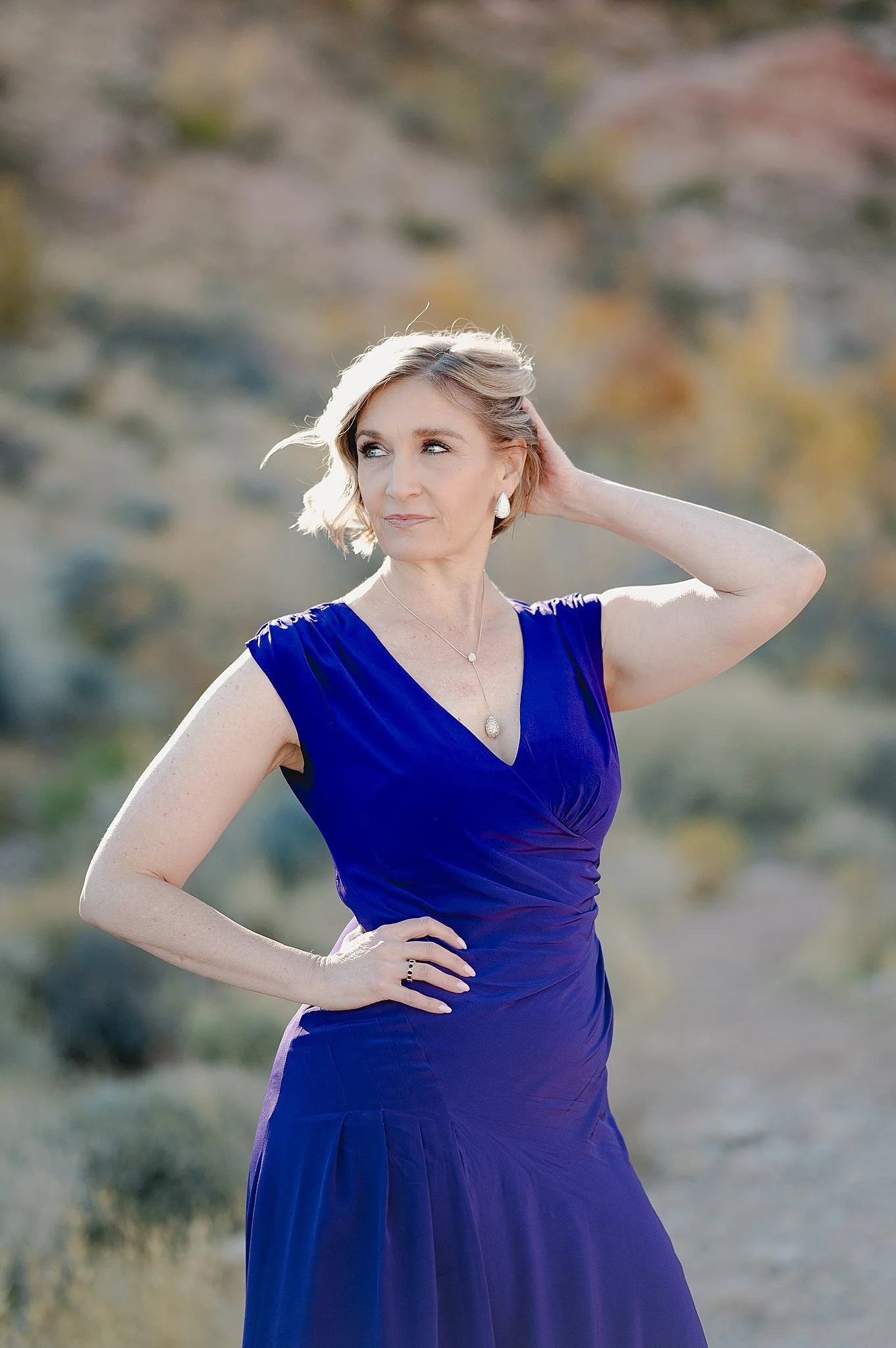 A woman in a royal blue dress stands outdoors with a blurred natural landscape in the background, posing with one hand on her hip and the other touching her hair, looking to the side
