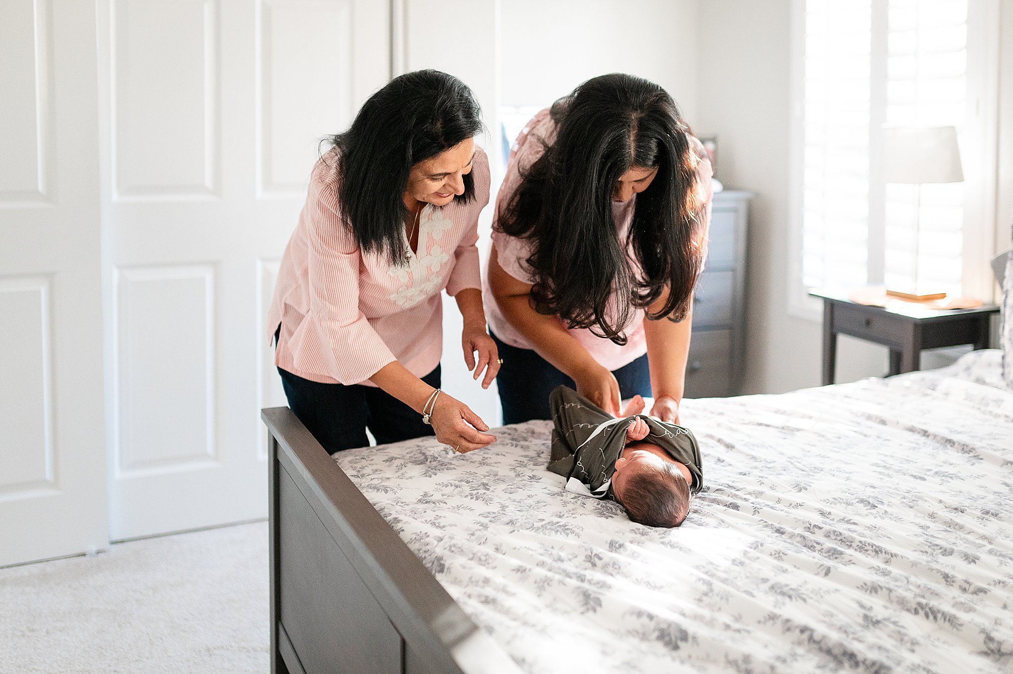 A mother and grandmother, swaddling a newborn baby boy, during an in-home newborn session.