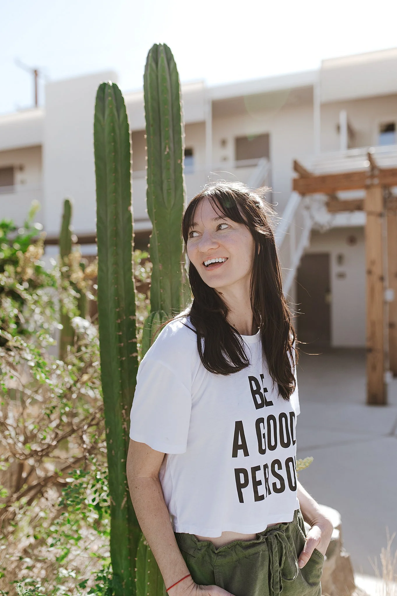 Woman with dark hair wearing a white T-shirt that says 'Be a good person,' standing outdoors near a tall cactus plant, smiling and looking to the side with residential buildings in the background.