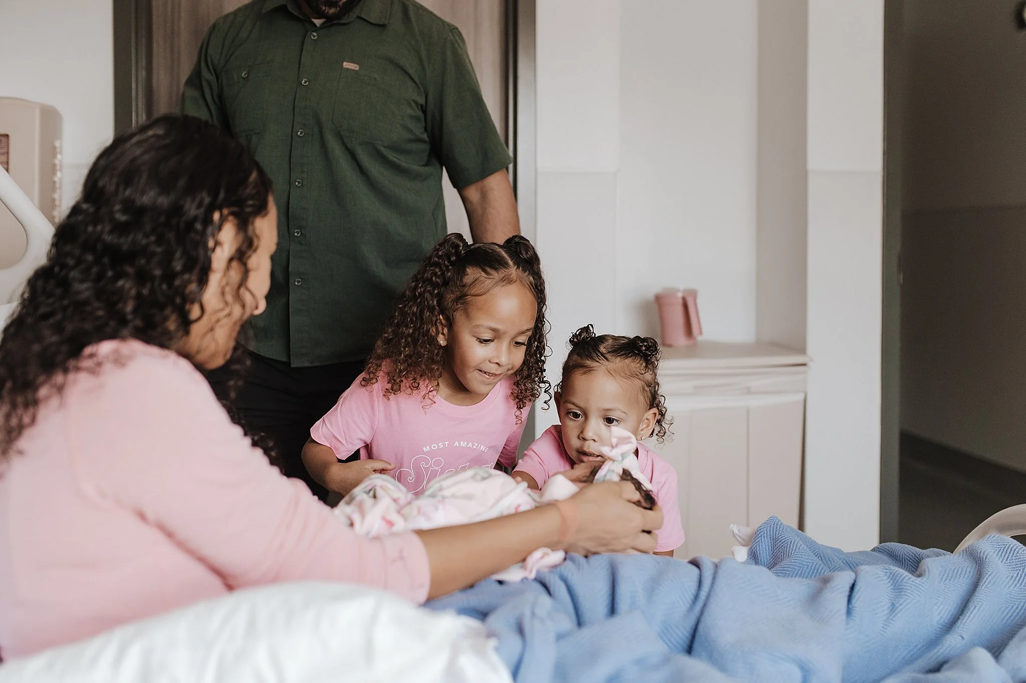 A woman in a pink shirt holds a newborn baby wrapped in a blanket, with two young girls in pink shirts looking on, all gathered around the baby in a hospital room, in Summerlin, Las Vegas.