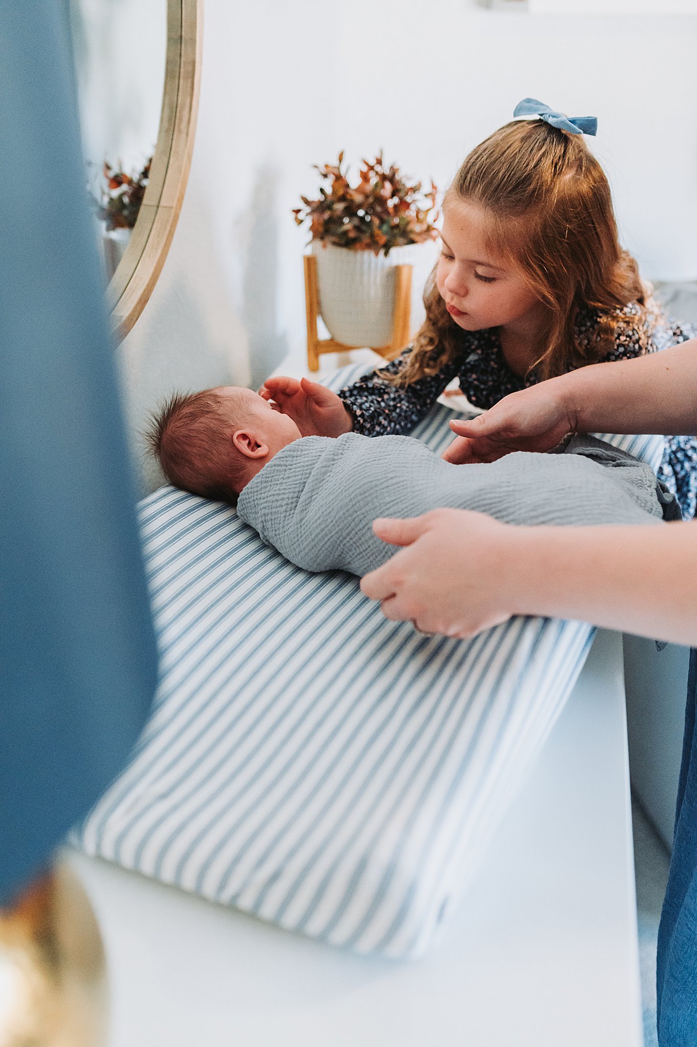 A toddler big sister looking on as her mom swaddles her baby brother, in a blue and white nursery.