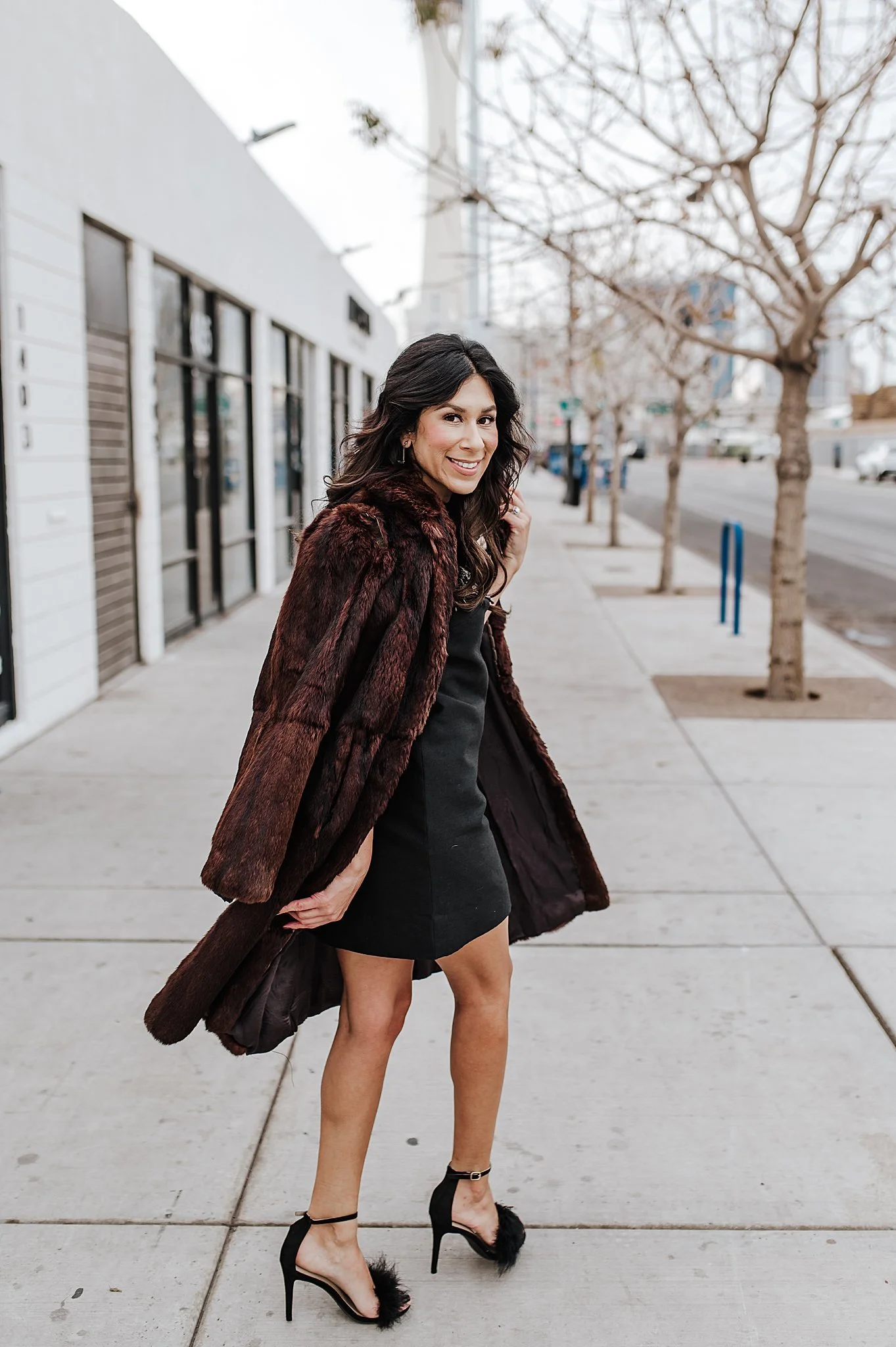 A woman with long dark wavy hair is standing on a city sidewalk, smiling at the camera. She is wearing a black dress, black high heels with furry details, and an open dark brown fur coat. In the background, there are leafless trees, storefronts, and
