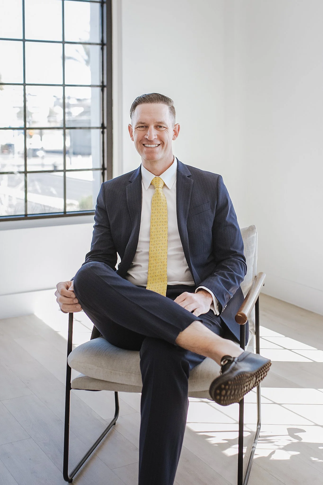 A man in a navy suit and yellow tie sitting on a chair, smiling at the camera in a bright room with large windows.