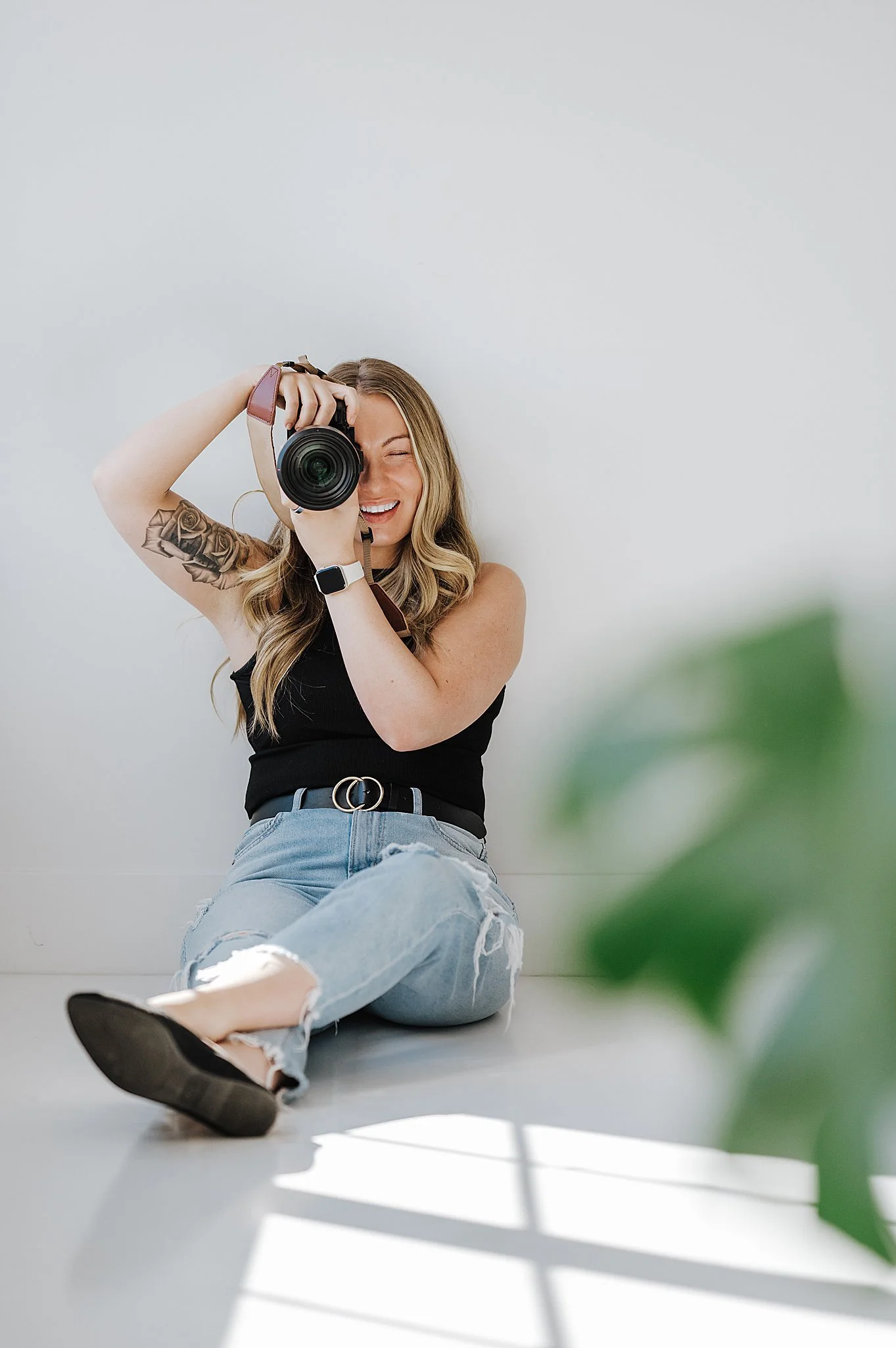 A young woman with wavy hair, tattoos, and a black tank top, sitting on the floor with legs crossed, taking a selfie with a camera, smiling with her eyes closed, next to a green plant and sunlight streaming through a window.