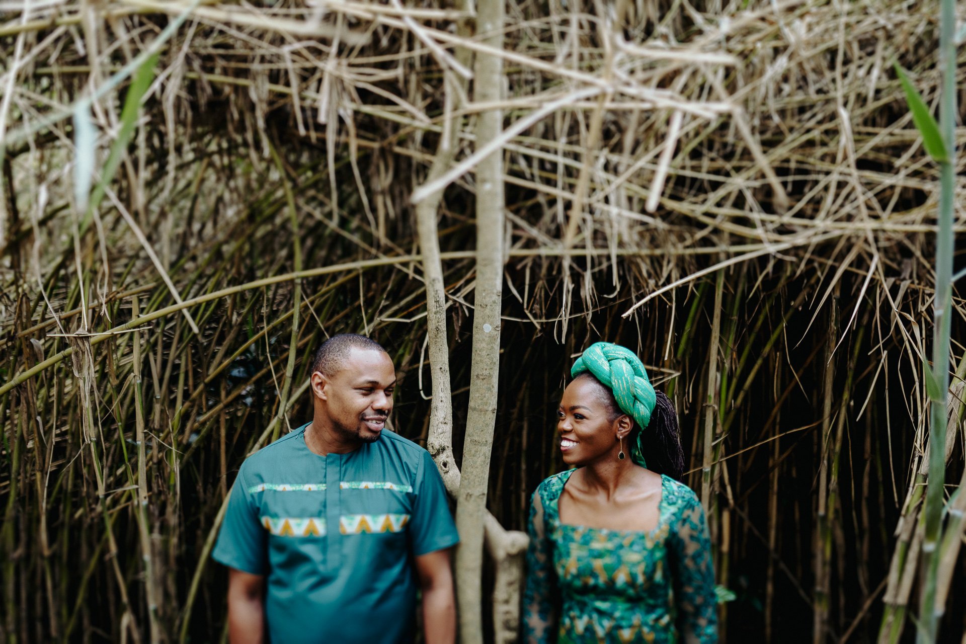 A man and woman are standing in front of tall dry reeds or grass, smiling and looking at each other. The woman is wearing a green headwrap and a patterned dress, while the man is dressed in a blue shirt with patterned details.