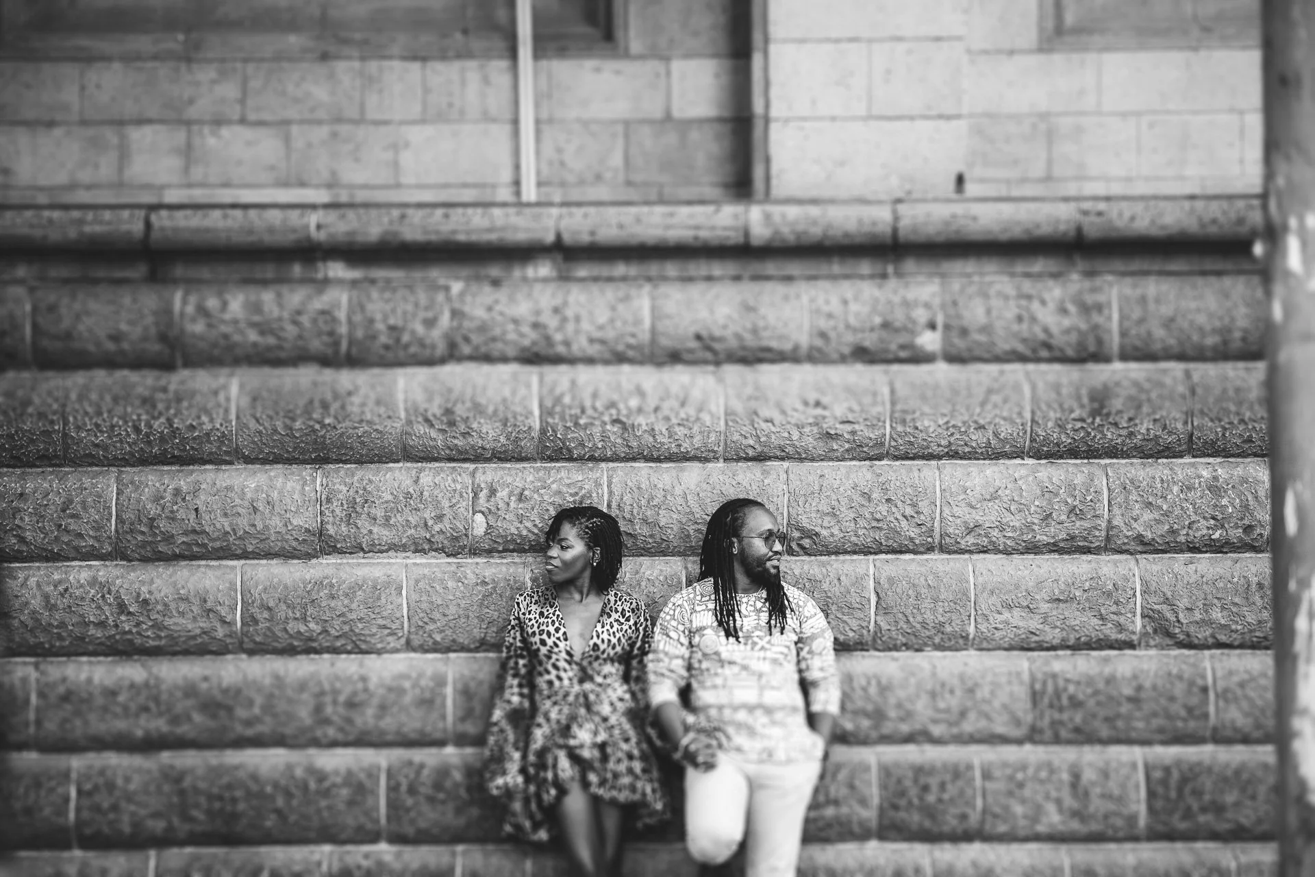 A black and white photo of a man and woman sitting on stone steps, holding hands. The woman is wearing a patterned dress and the man is wearing a patterned shirt and pants. They are looking in opposite directions.
