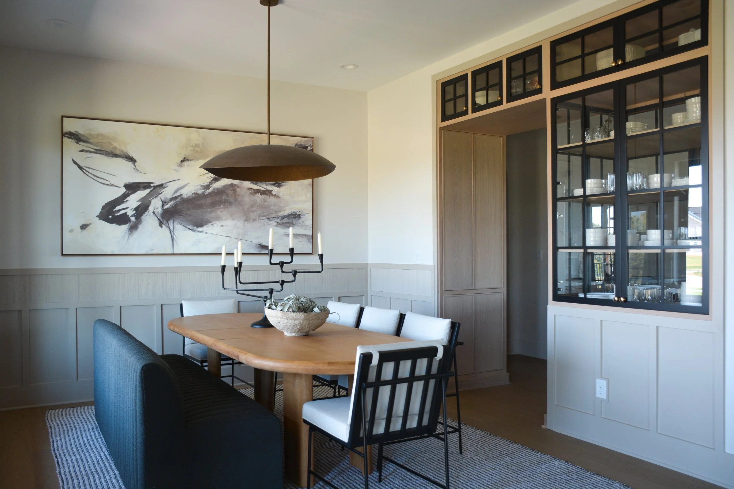 Dining room featuring a wood dining table, built-in cabinetry with glass doors, oversized artwork, and layered lighting in a transitional interior.