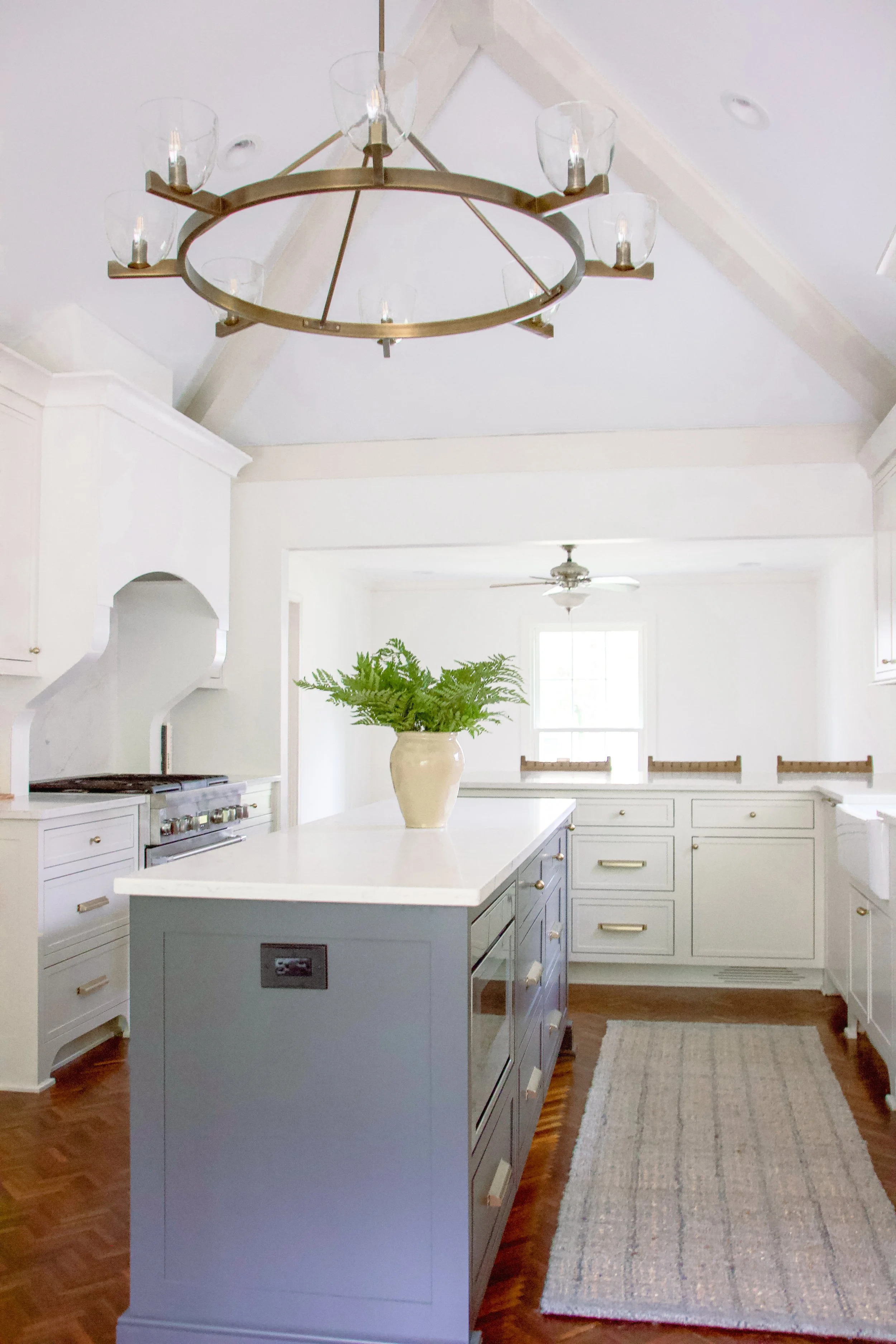 Studio Nine Design’s bright, transitional kitchen pairs white shaker cabinetry with a blue-gray island and quartz surfaces. Brass hardware and a ring chandelier add warmth while the long runner and wood floors ground the space.