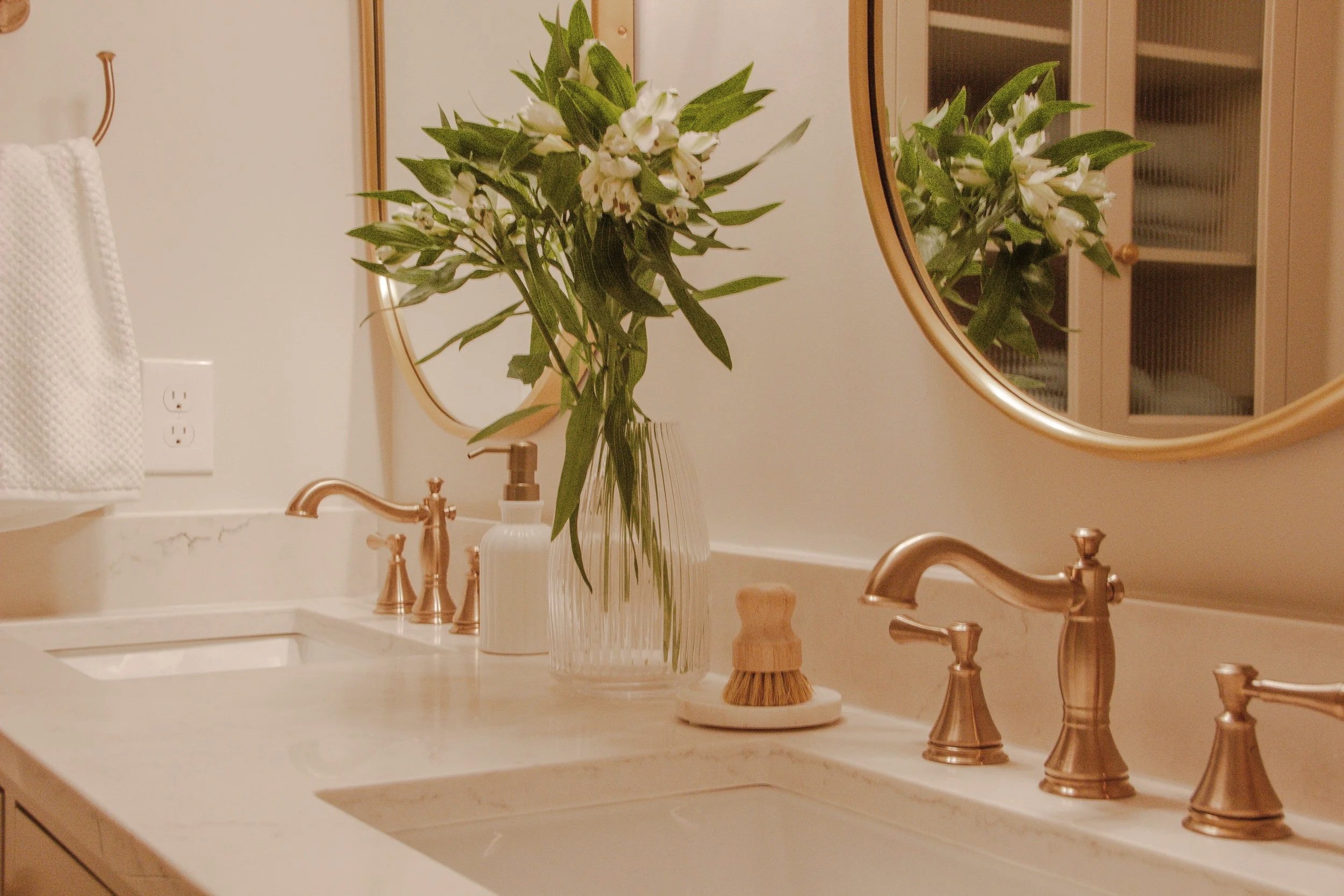 Stone vanity countertop with integrated sinks, warm brass faucets, rounded mirrors, and simple accessories arranged with restraint.