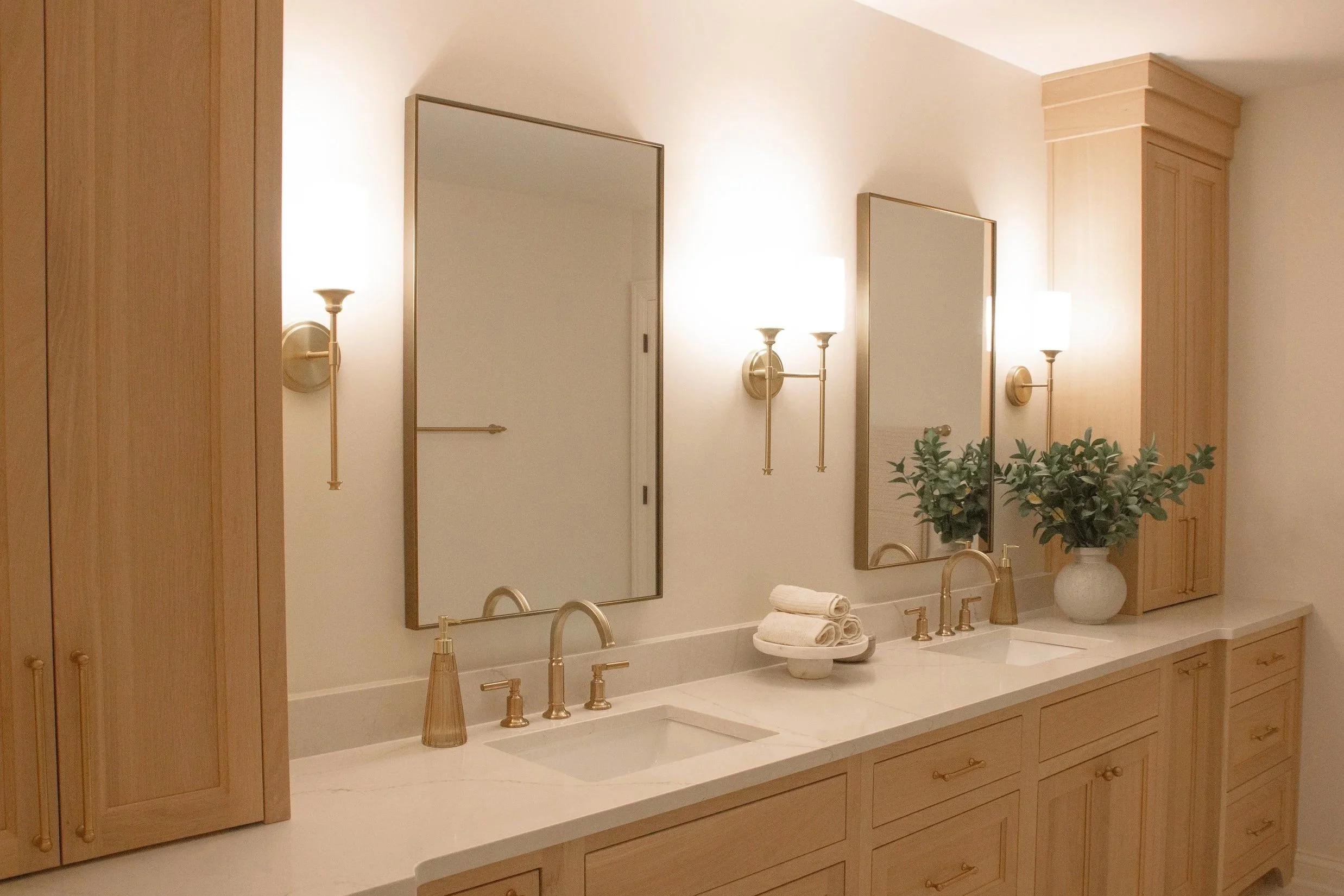 Light wood double vanity with stone countertop, brass fixtures, and balanced lighting creating a calm, spa-inspired bathroom environment.