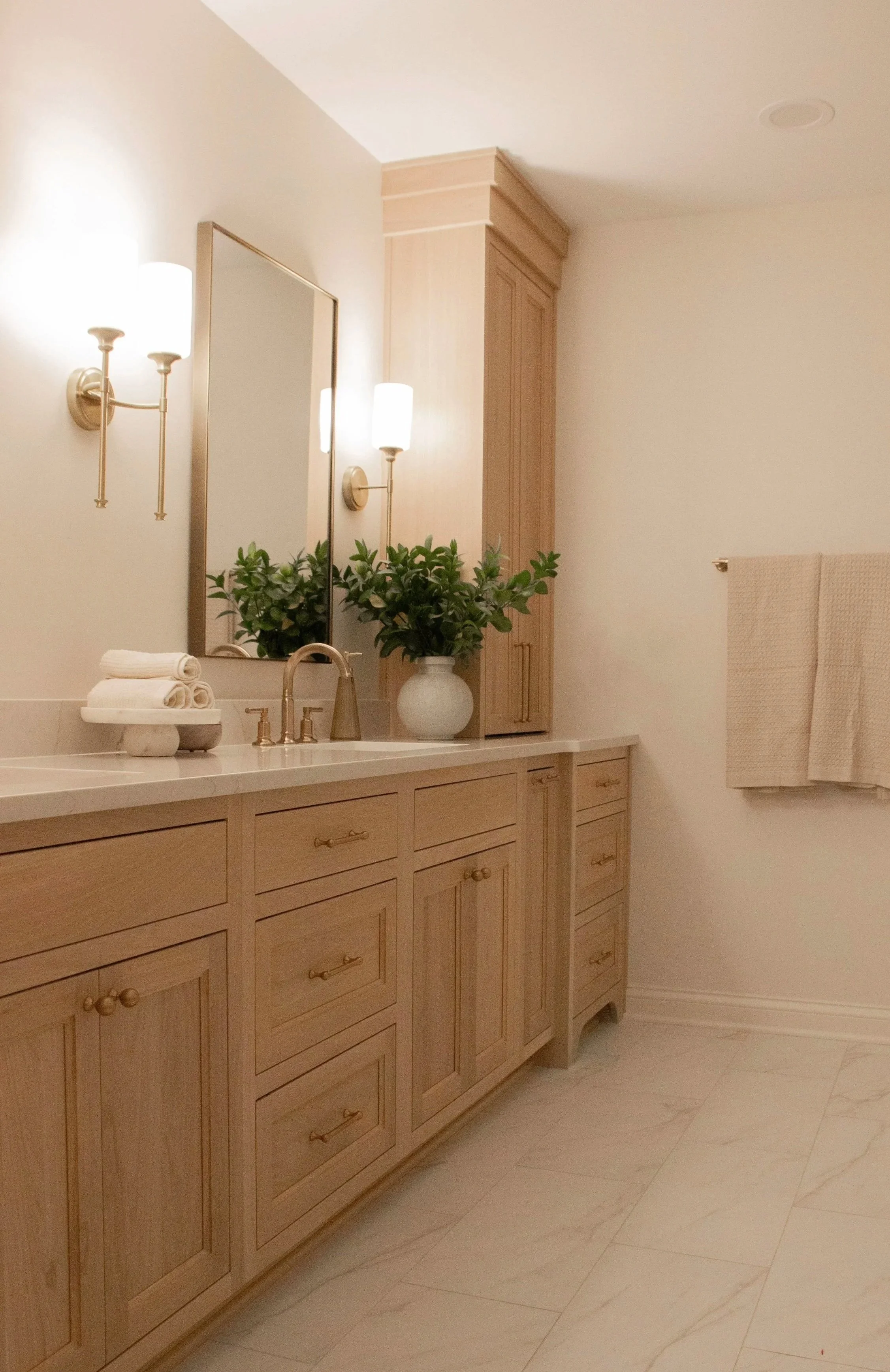 Light wood double vanity with stone countertop, brass fixtures, and wall sconces creating a calm, spa-inspired vanity area.
