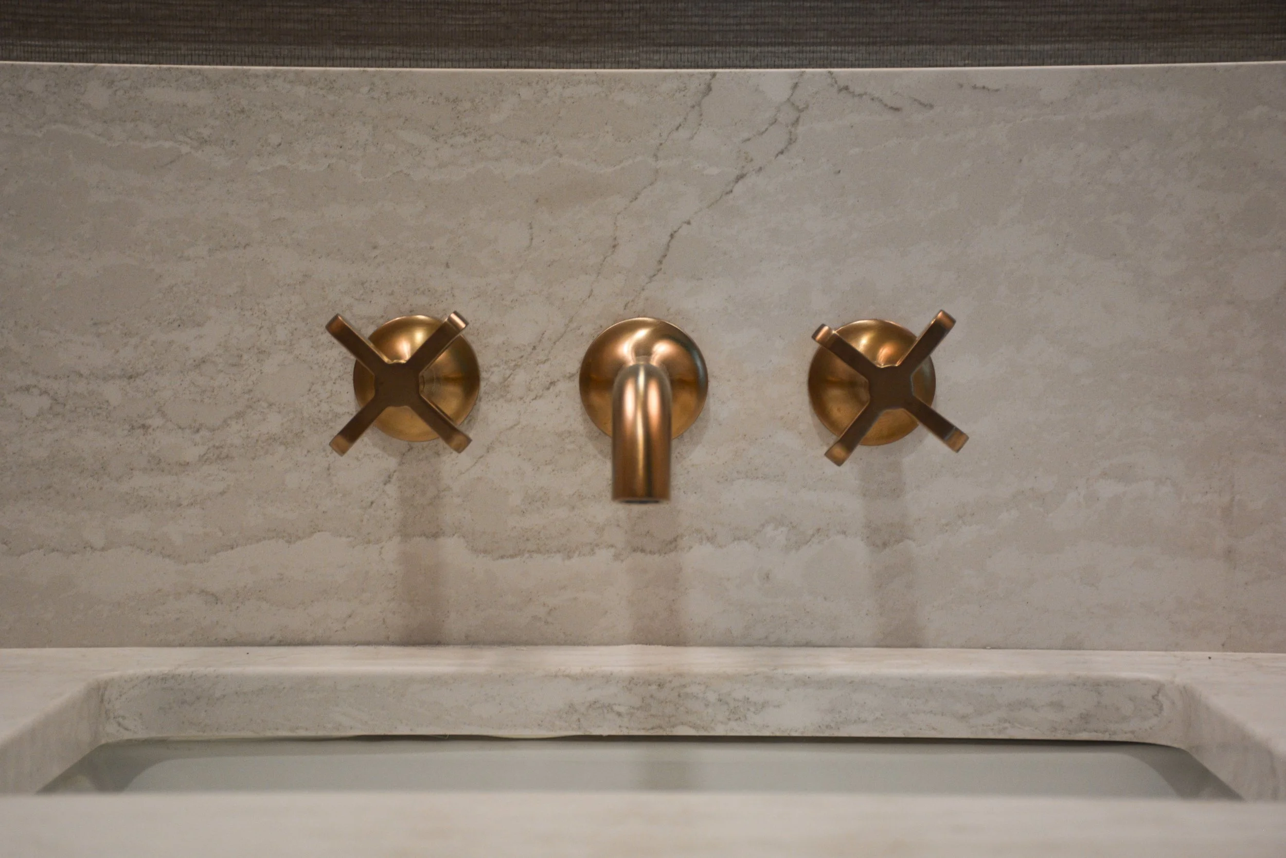 Wall-mounted brass faucet and cross handles set against a stone backsplash, emphasizing symmetry, material contrast, and restrained detailing.
