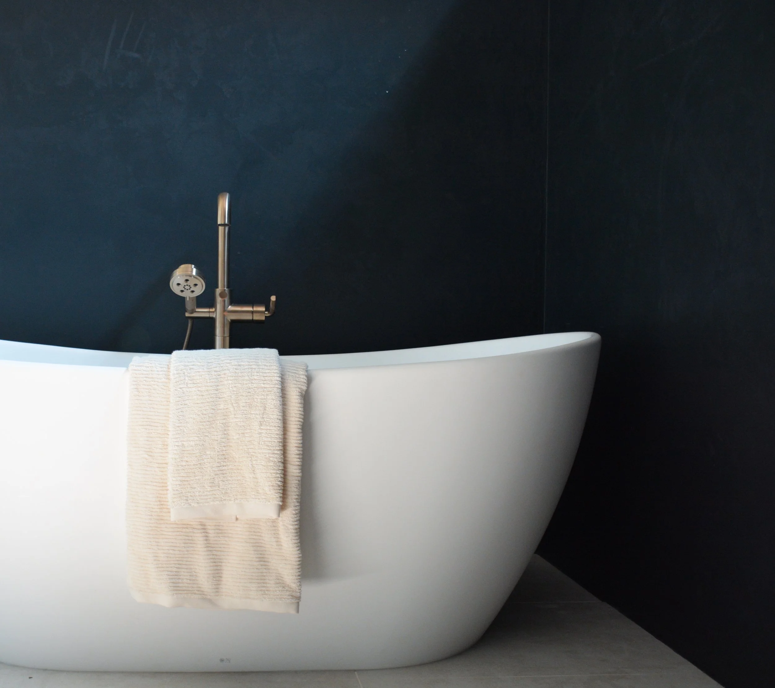 Freestanding white soaking tub with a brushed brass floor-mounted faucet positioned against a dark textured accent wall in a renovated primary bathroom.