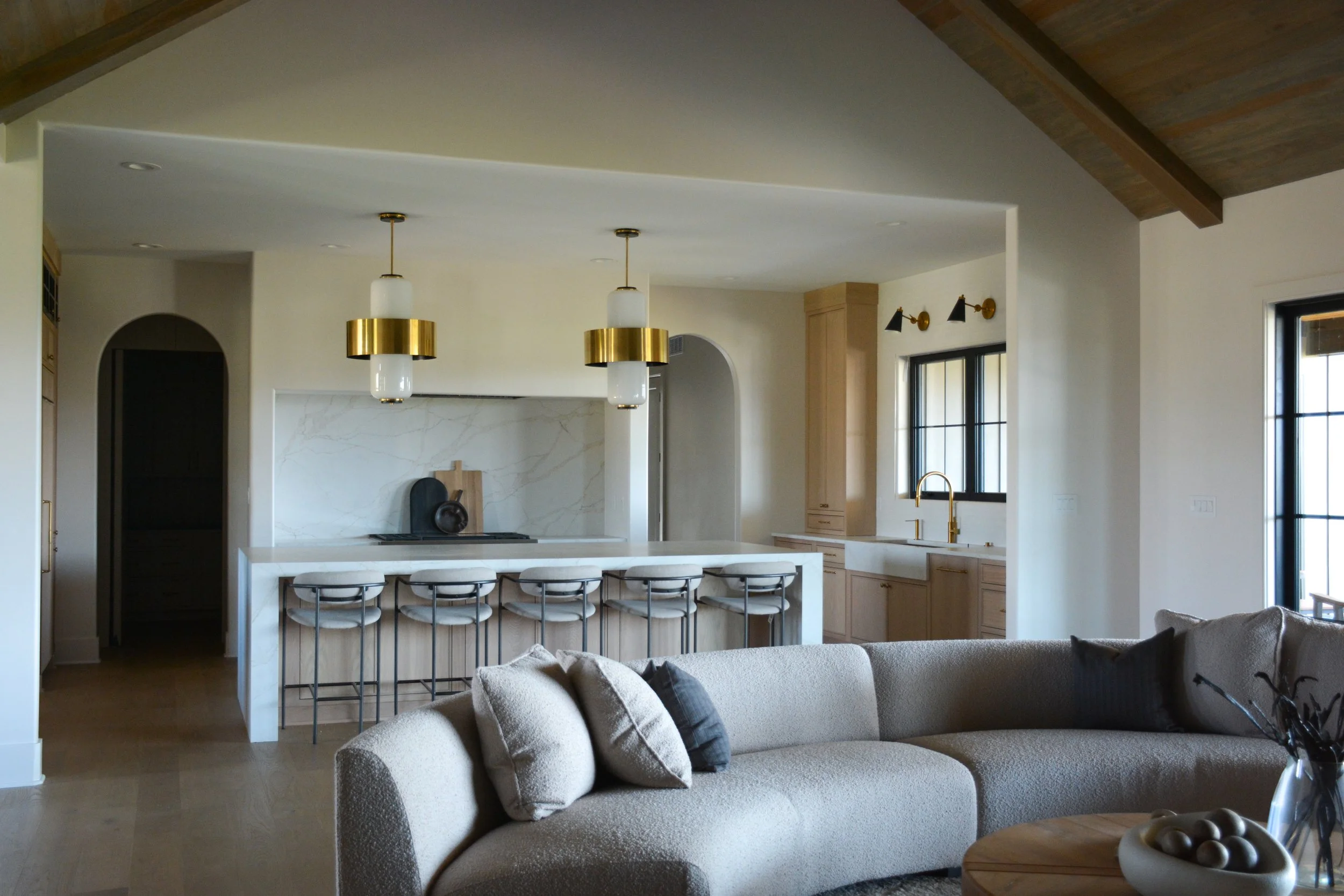 Open-concept kitchen and living room featuring a curved sofa, waterfall island, warm wood accents, brass lighting, and vaulted ceilings designed as part of a full-scale home renovation.