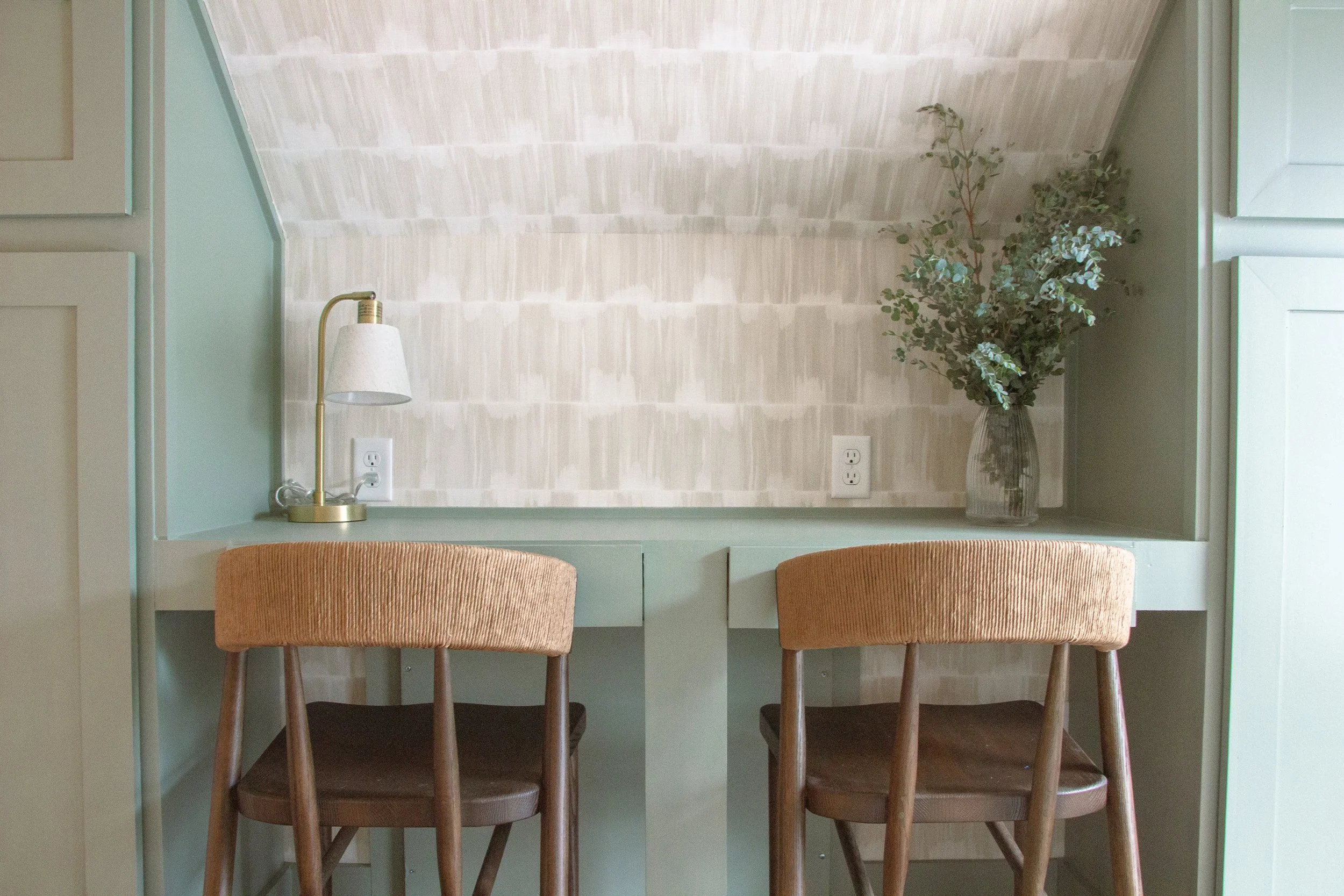 Built-in workspace featuring dual desks, patterned wallpaper, integrated cabinetry, and woven chairs designed as part of a full-scale home renovation.