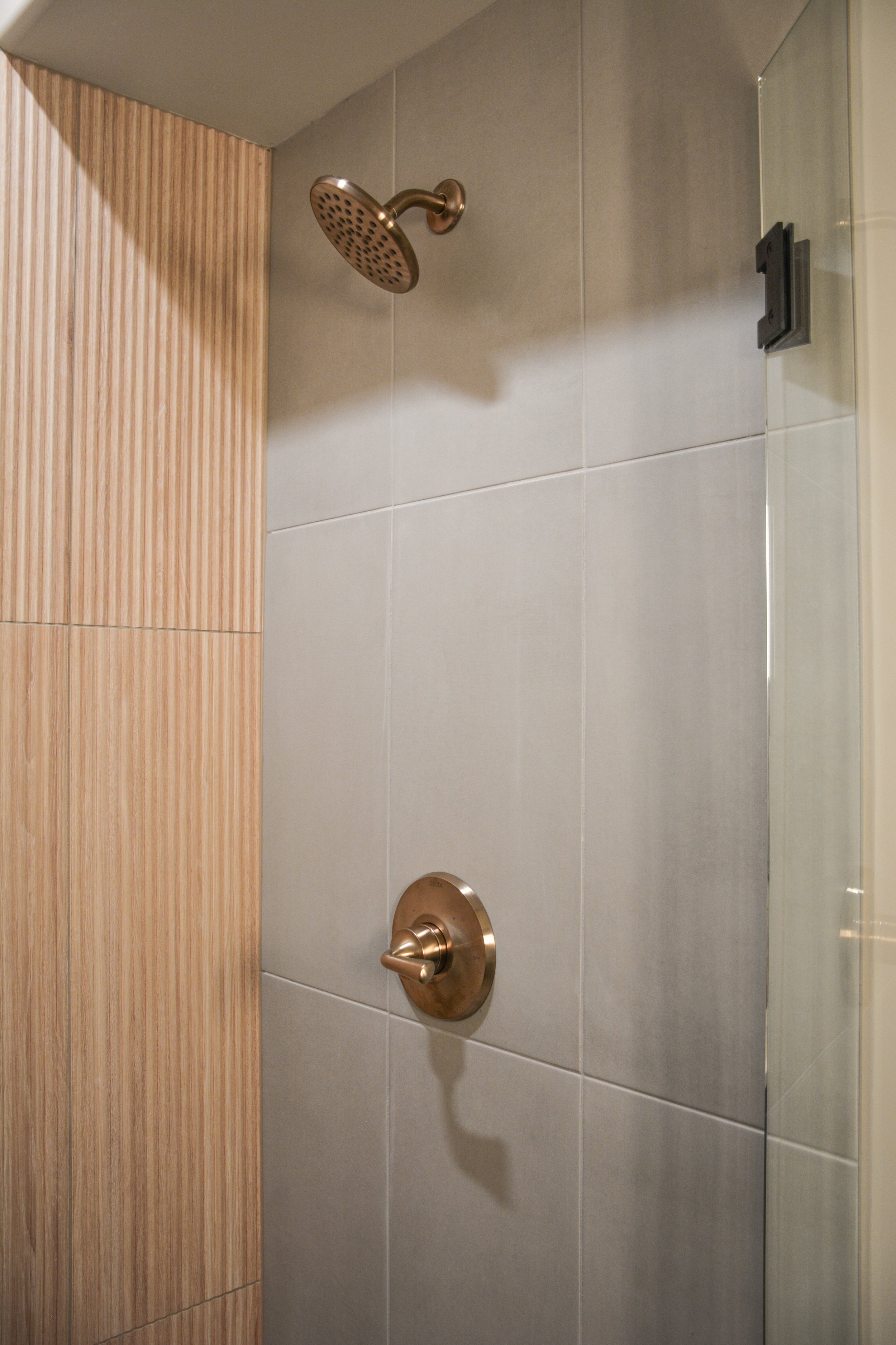Shower wall with light stone tile, warm metal fixtures, and vertical wood detailing demonstrating how lighting affects surface color.