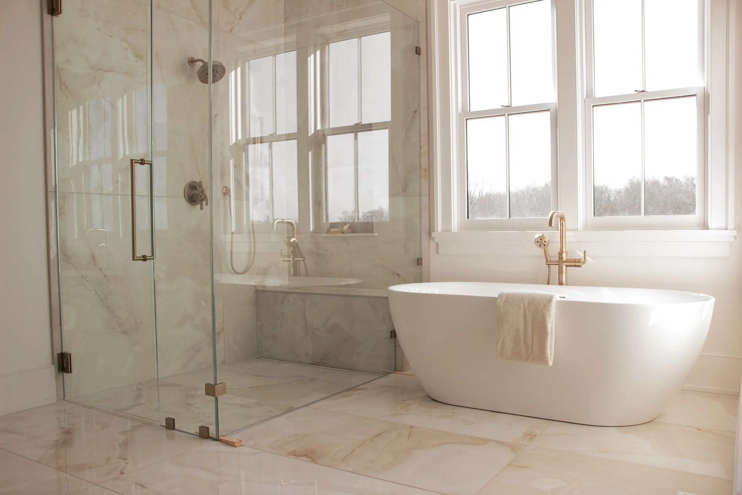 Primary bathroom featuring a freestanding soaking tub, glass-enclosed shower, marble-look tile, brass fixtures, and large windows designed as part of a full-scale home renovation.