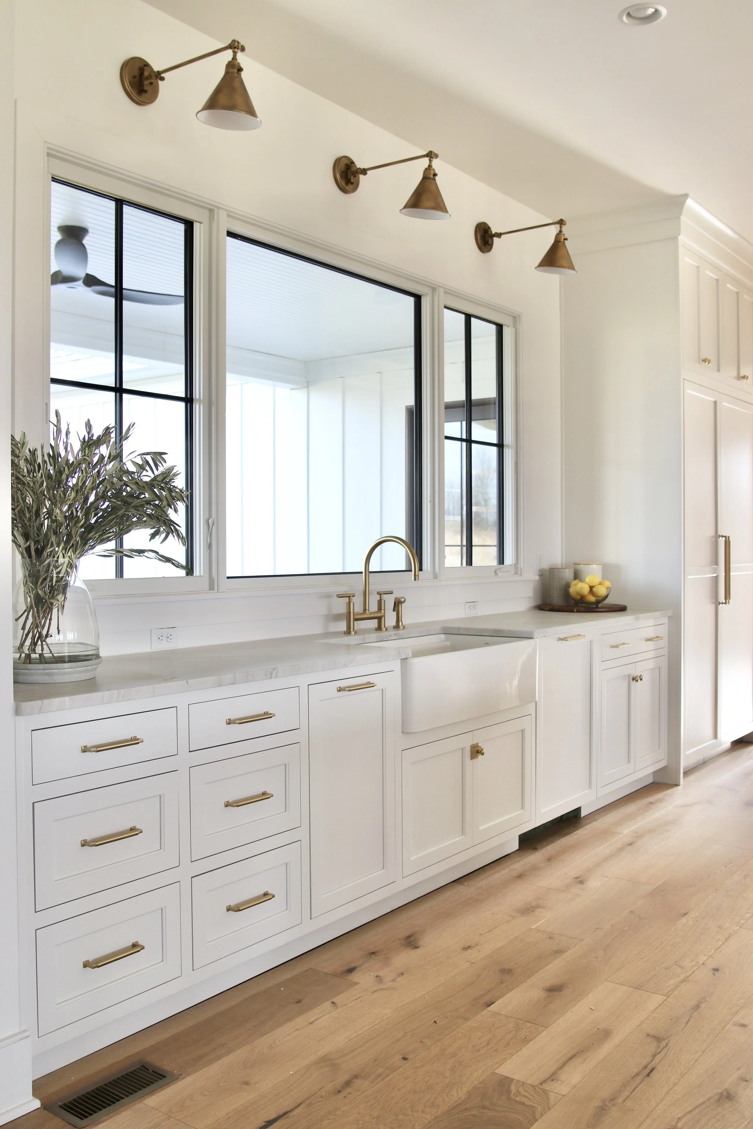 This bright, open kitchen design features crisp white cabinetry with gold brass handles and fixtures. A large picture window above the farmhouse sink brings in natural light. The window is framed by five brass sconces, which add warmth and contrast t