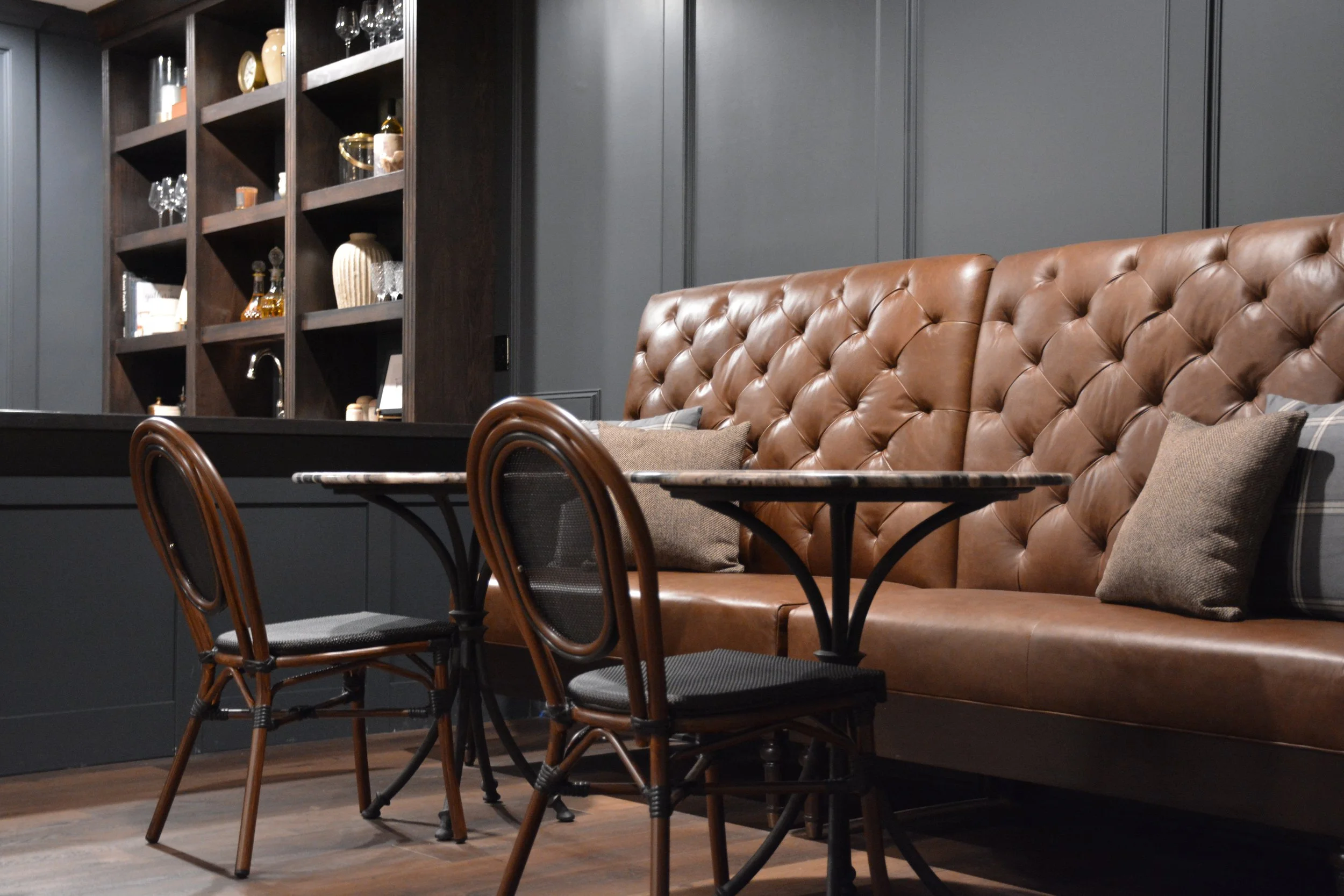 Tufted leather banquette with marble café tables and dark wood built-in bar shelving in a finished basement.