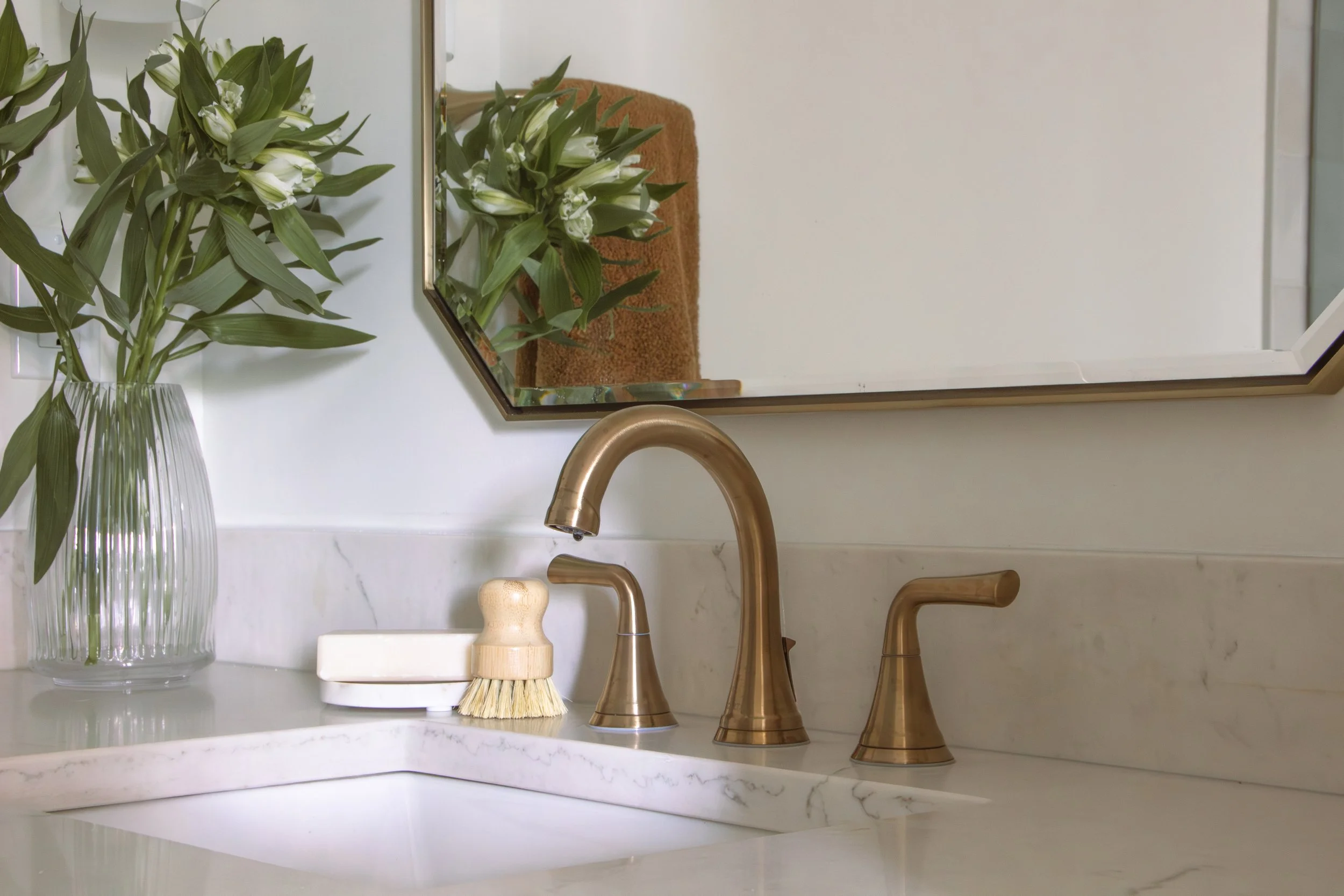 Bathroom vanity featuring brass fixtures, a marble countertop, a geometric mirror, and fresh greenery designed as part of a full-scale home renovation.