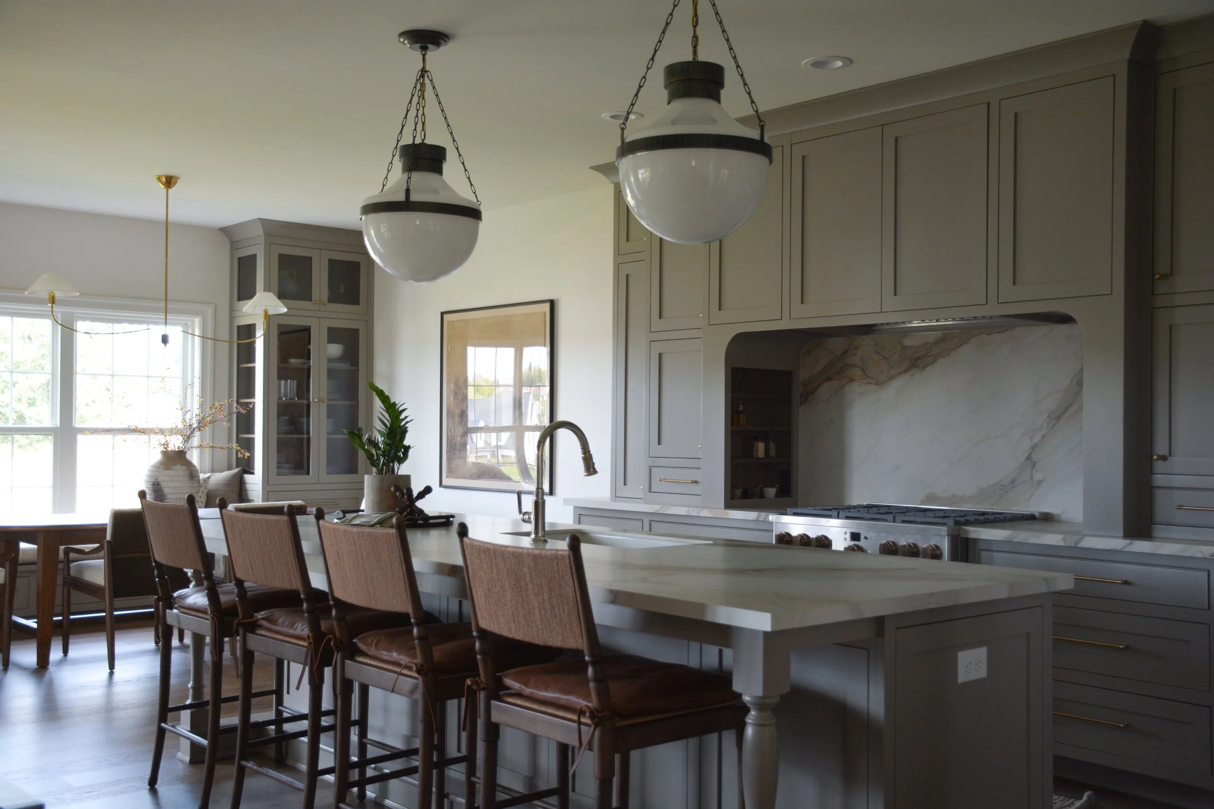 This kitchen showcases a sophisticated, moody palette with full-height gray cabinetry and a dramatic marble slab backsplash behind the cooking range. A large island with a waterfall-edge countertop includes an undermount sink and four wood-framed lea