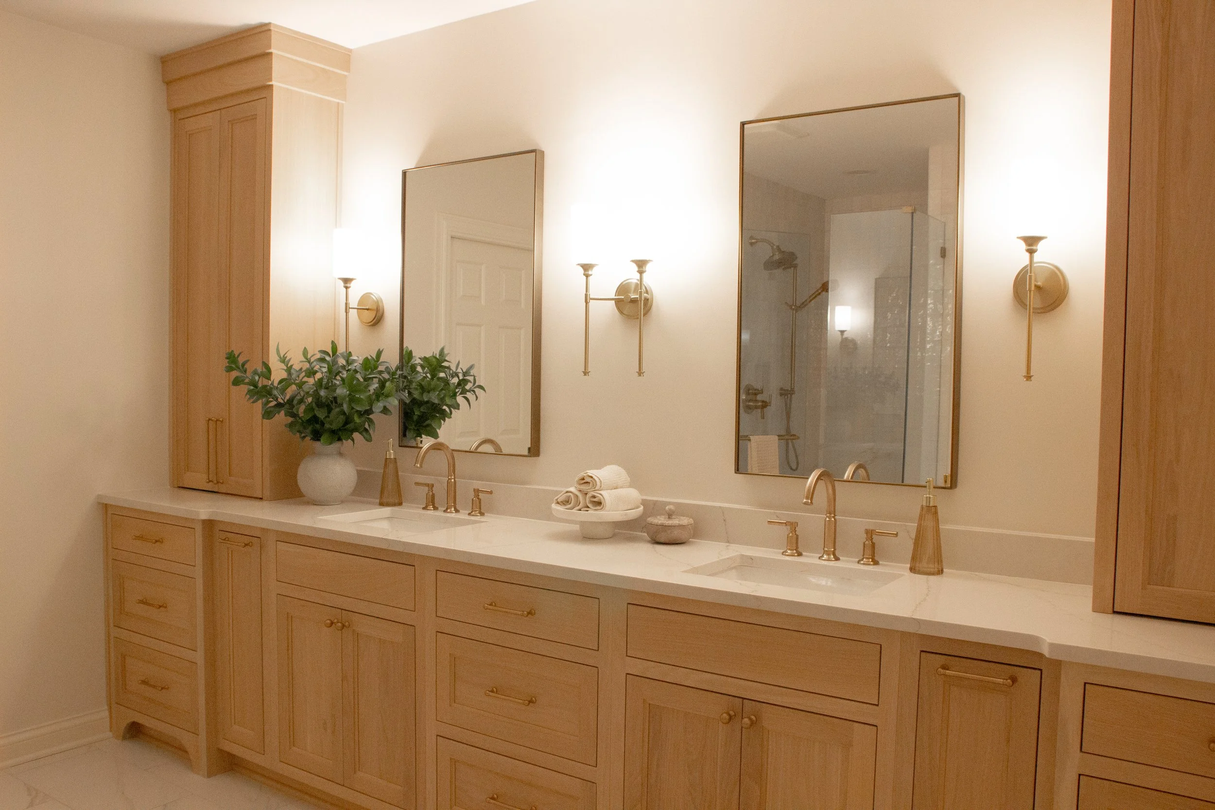Light wood double vanity with integrated storage, stone countertop, brass fixtures, and balanced wall lighting creating a calm, spa-inspired environment.