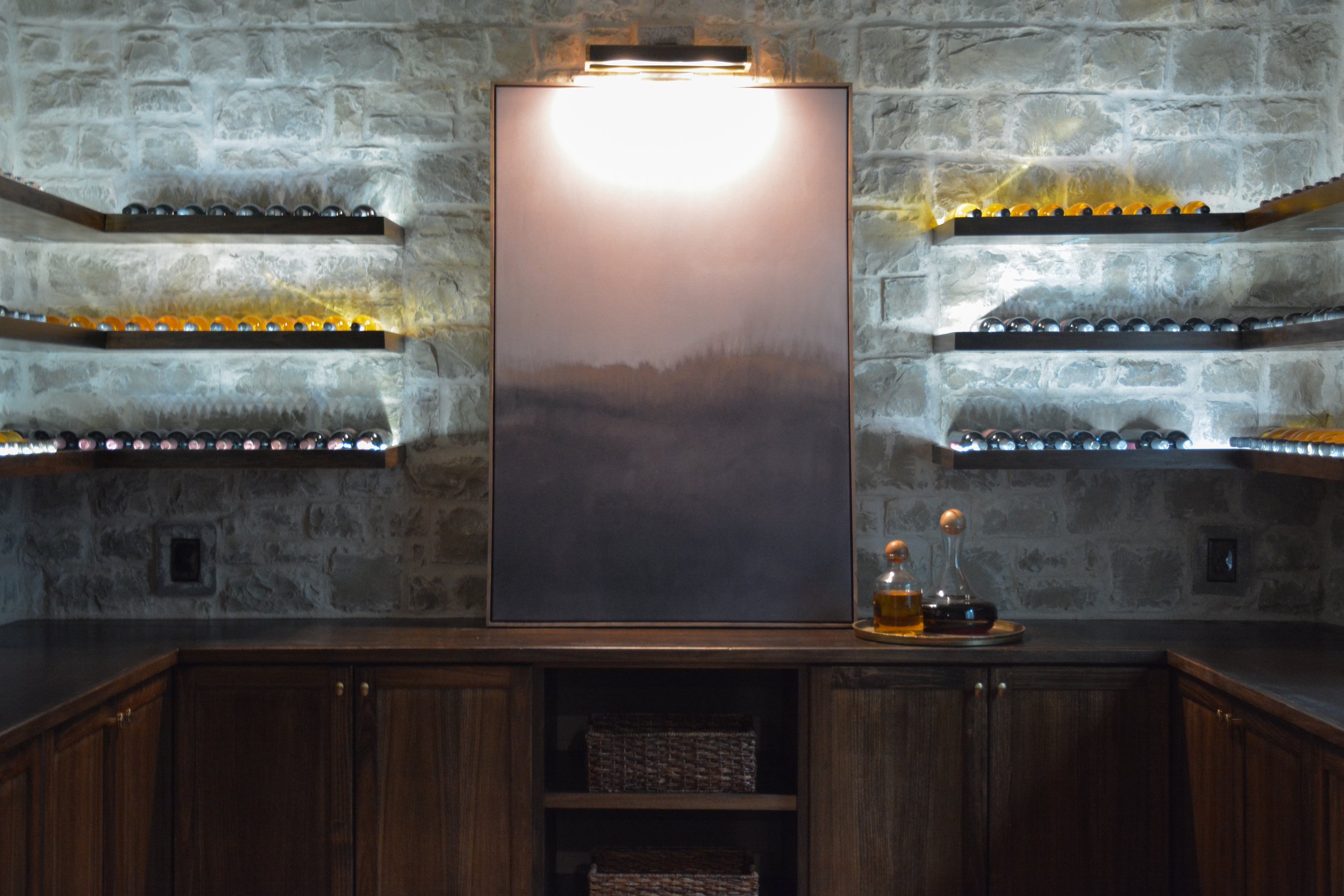 Stylish basement bar wall with illuminated shelving, decorative bottles, and a central art piece framed by warm lighting, adding depth and character to the lower level.
