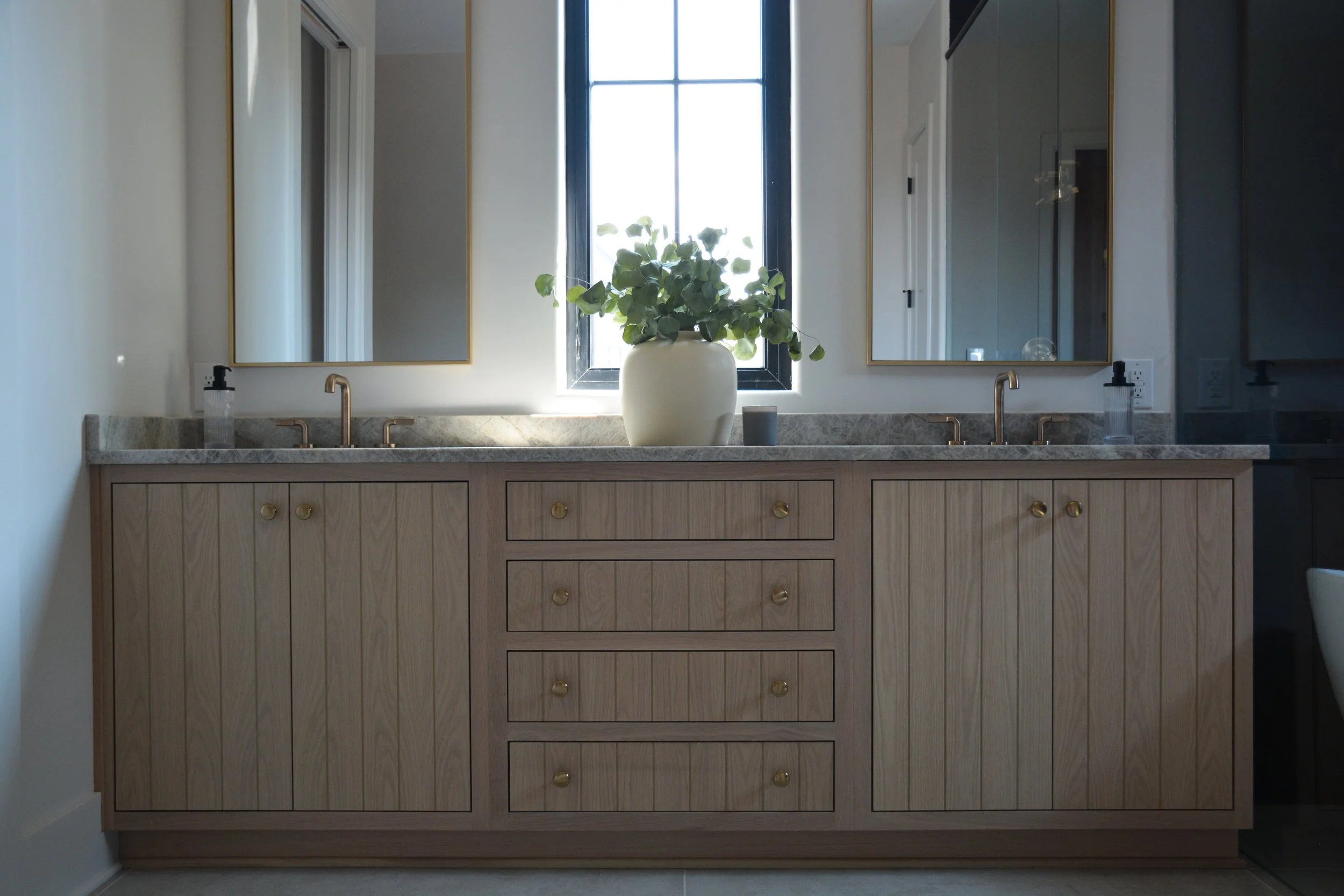 Wood vanity with integrated drawers beneath a stone countertop, flanked by mirrors and centered under a window that brings in natural light and airflow.