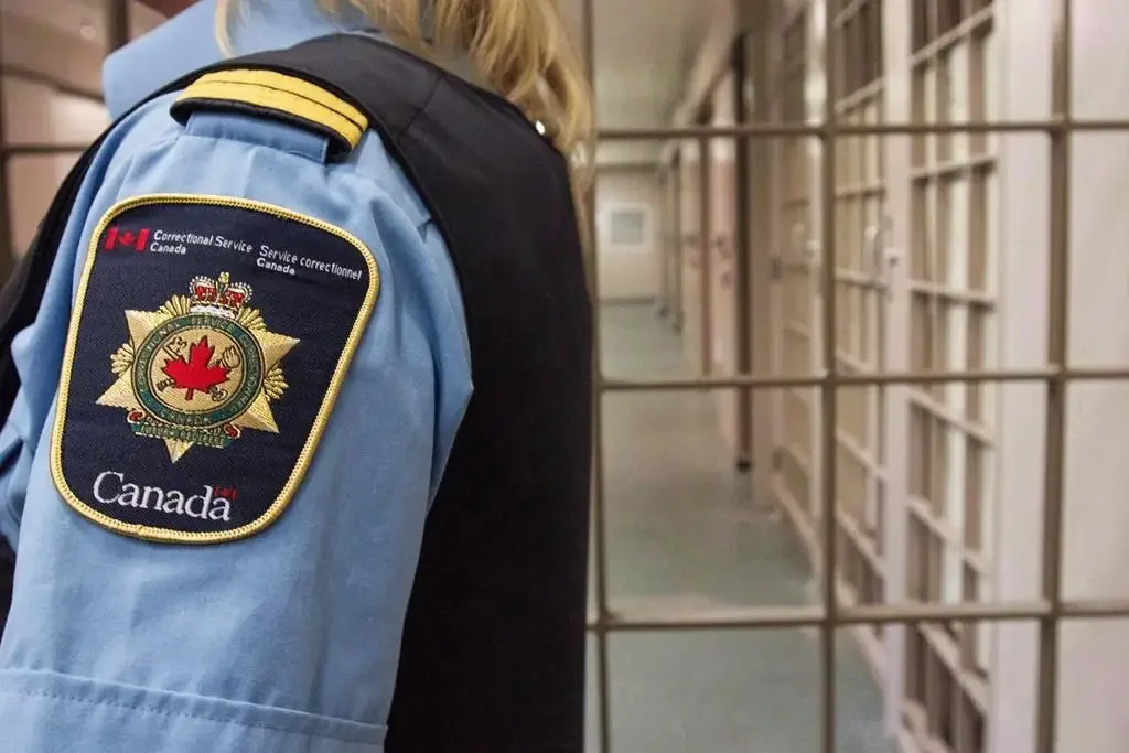 A Canadian correctional officer in uniform standing in a prison hallway with jail cells on the right.
