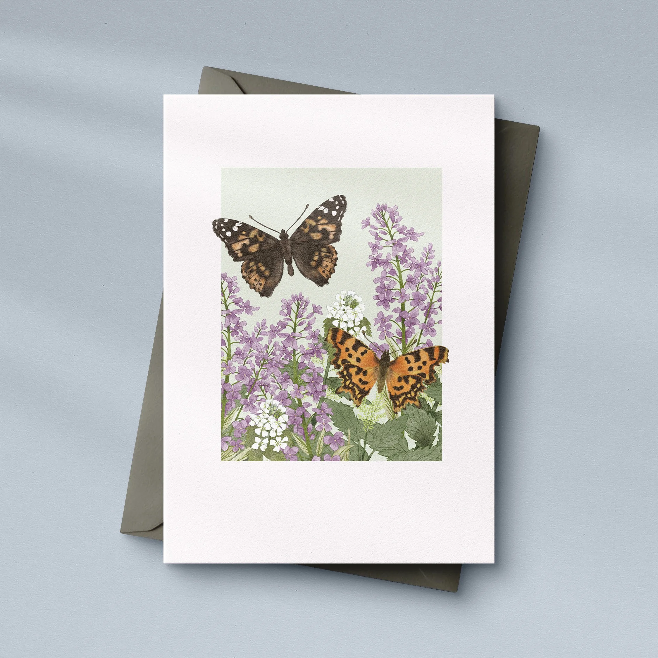 Greeting card with watercolour illustration of a painted lady butterfly and a comma butterfly flying in a garden.The card is sitting on a dark grey envelope.