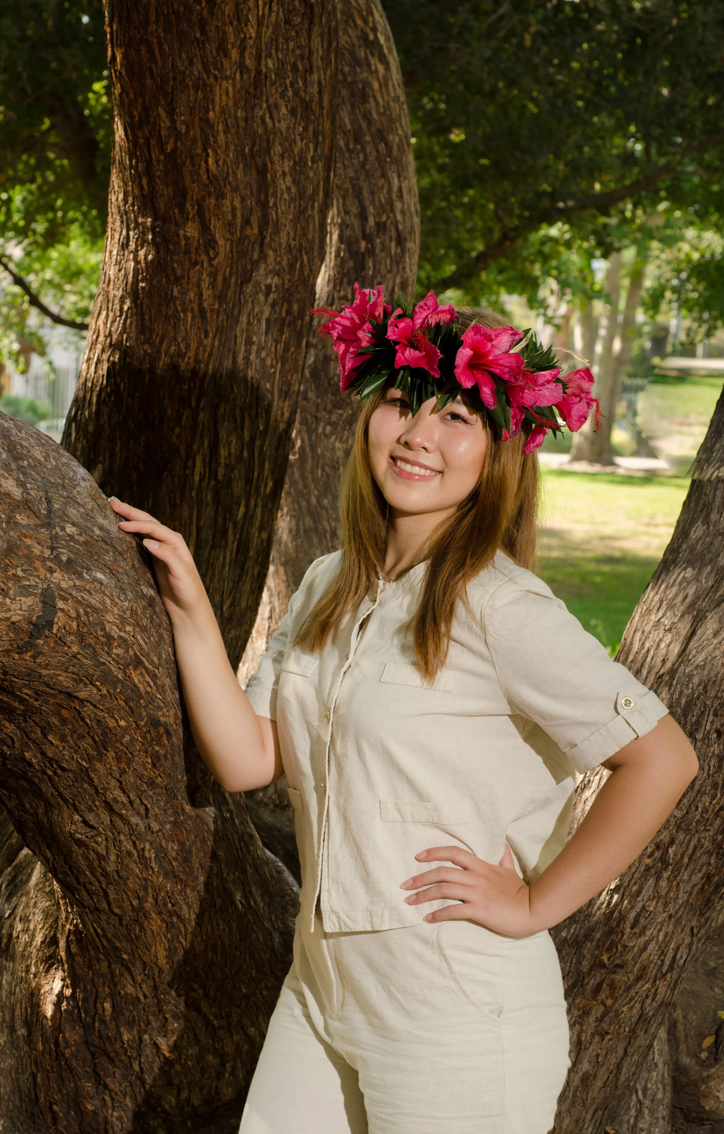 A woman standing next to a large tree, wearing a floral crown of pink flowers, smiling, with her right hand on her hip and her left hand touching the tree.