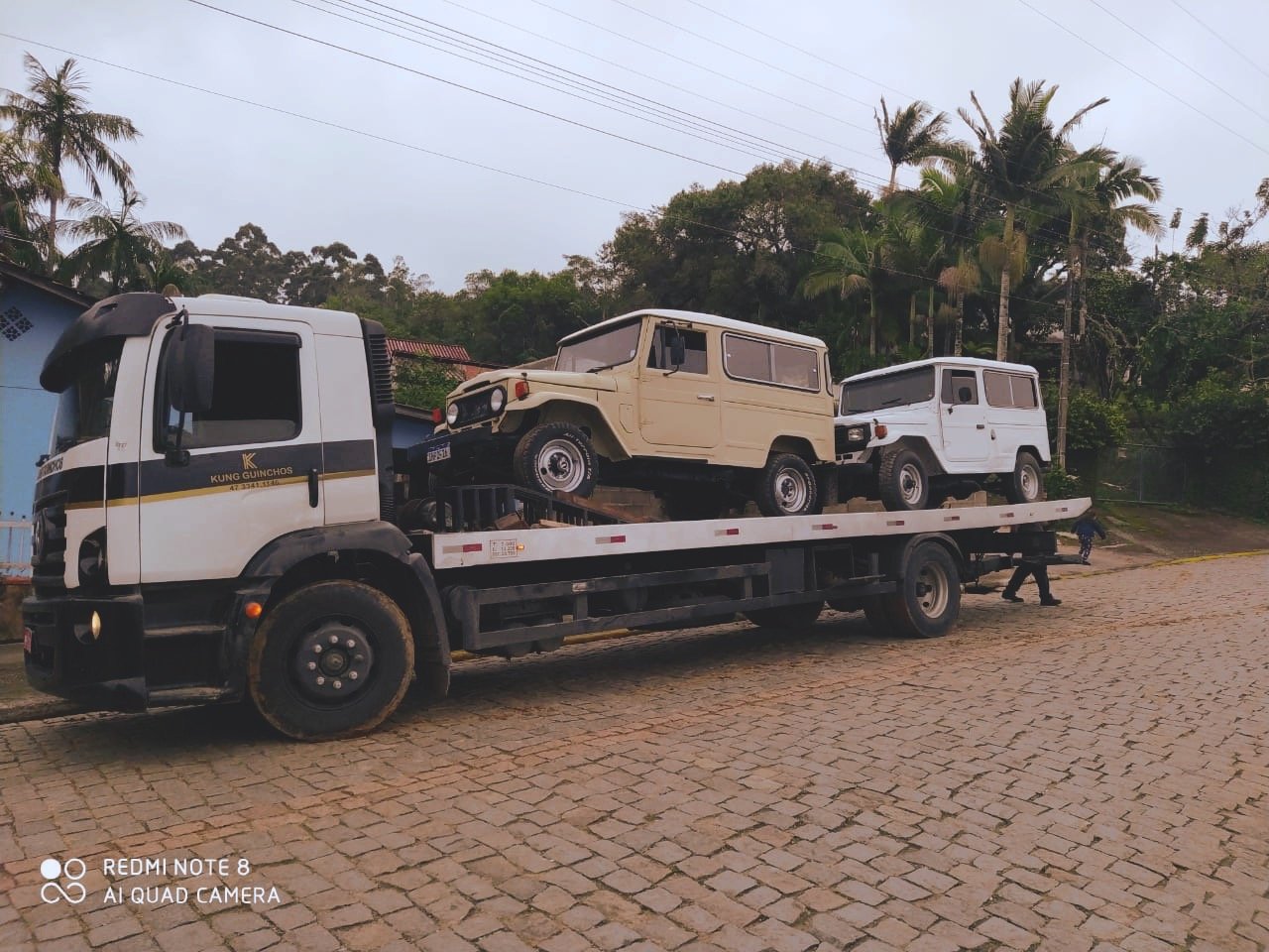 Flatbed truck transporting two vintage SUVs on a brick road with trees and houses in the background.
