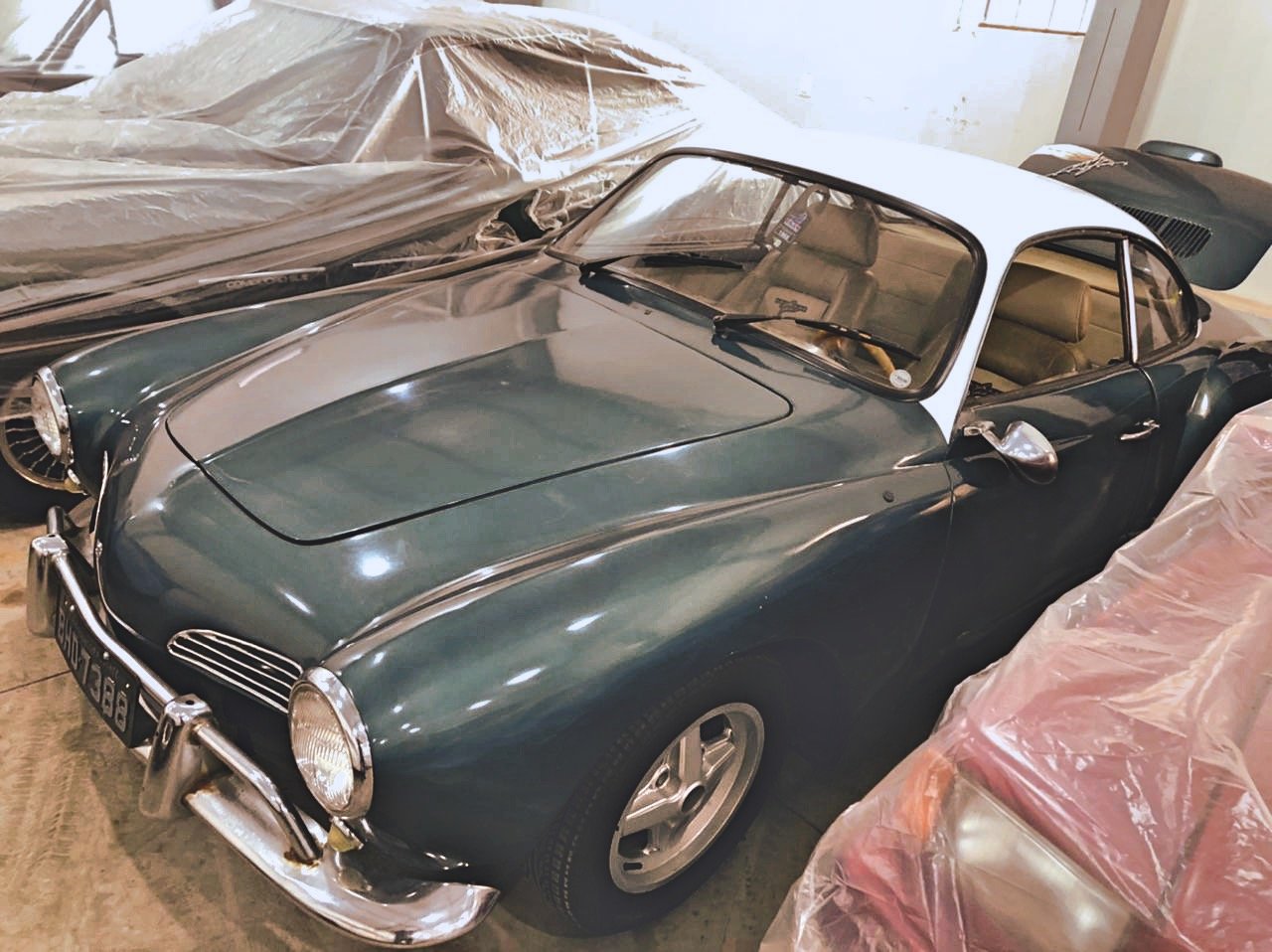 Vintage car collection, including a classic Volkswagen Karmann Ghia, partially covered with protective sheets in a storage space.