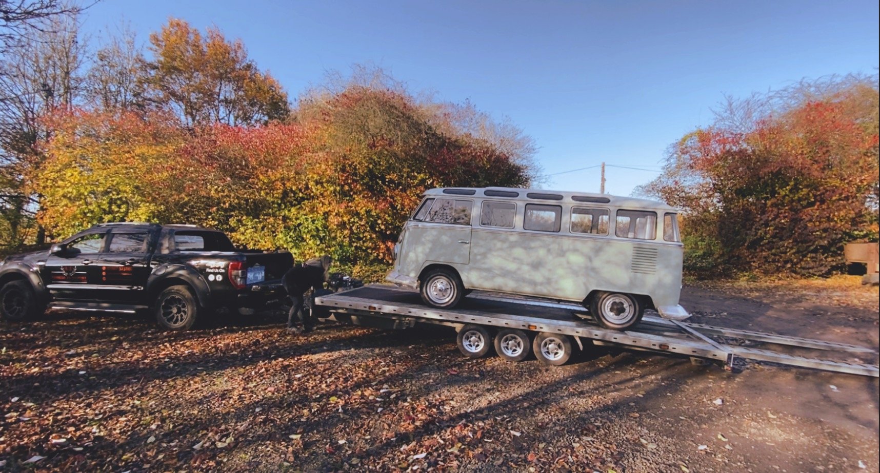 Black pickup truck towing a trailer with a light blue vintage van, surrounded by autumn foliage.