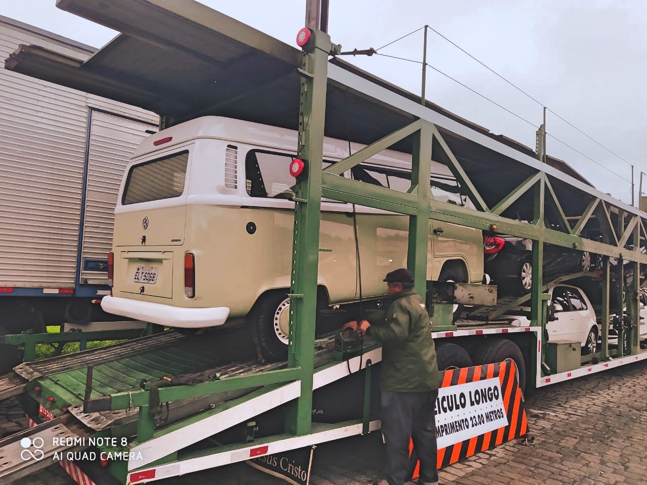 Transport truck carrying vehicles, including a VW van, multiple cars, person on right side.