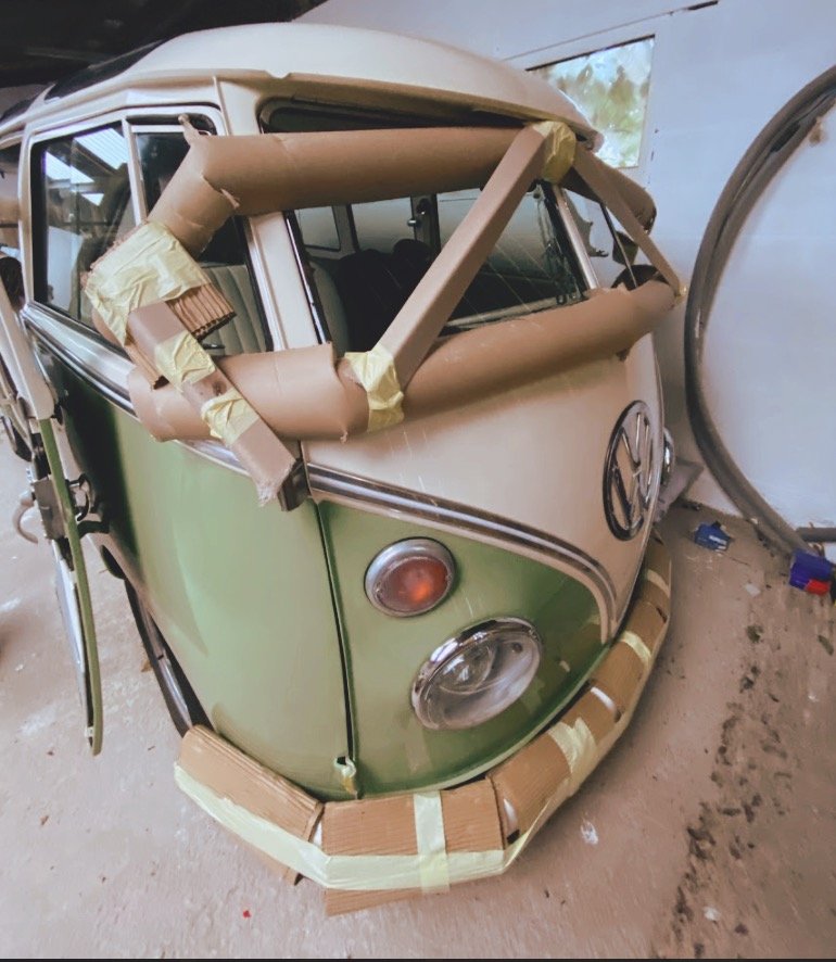 Vintage green and white Volkswagen van with cardboard and tape protective wrapping on bumpers and windows, parked indoors.
