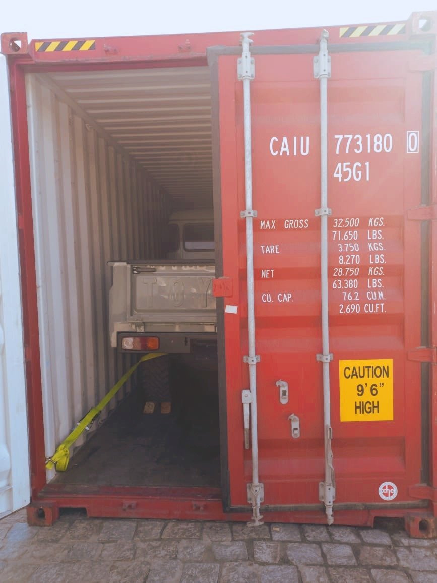 A truck loaded inside a red shipping container, secured with a yellow strap. The container's door is open, revealing specifications such as max gross weight and caution height.
