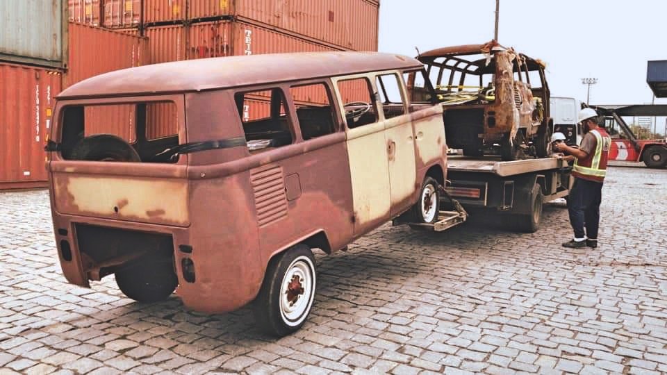 Rusty Volkswagen Type 2 van being loaded onto a tow truck in an industrial area with shipping containers and a worker nearby.