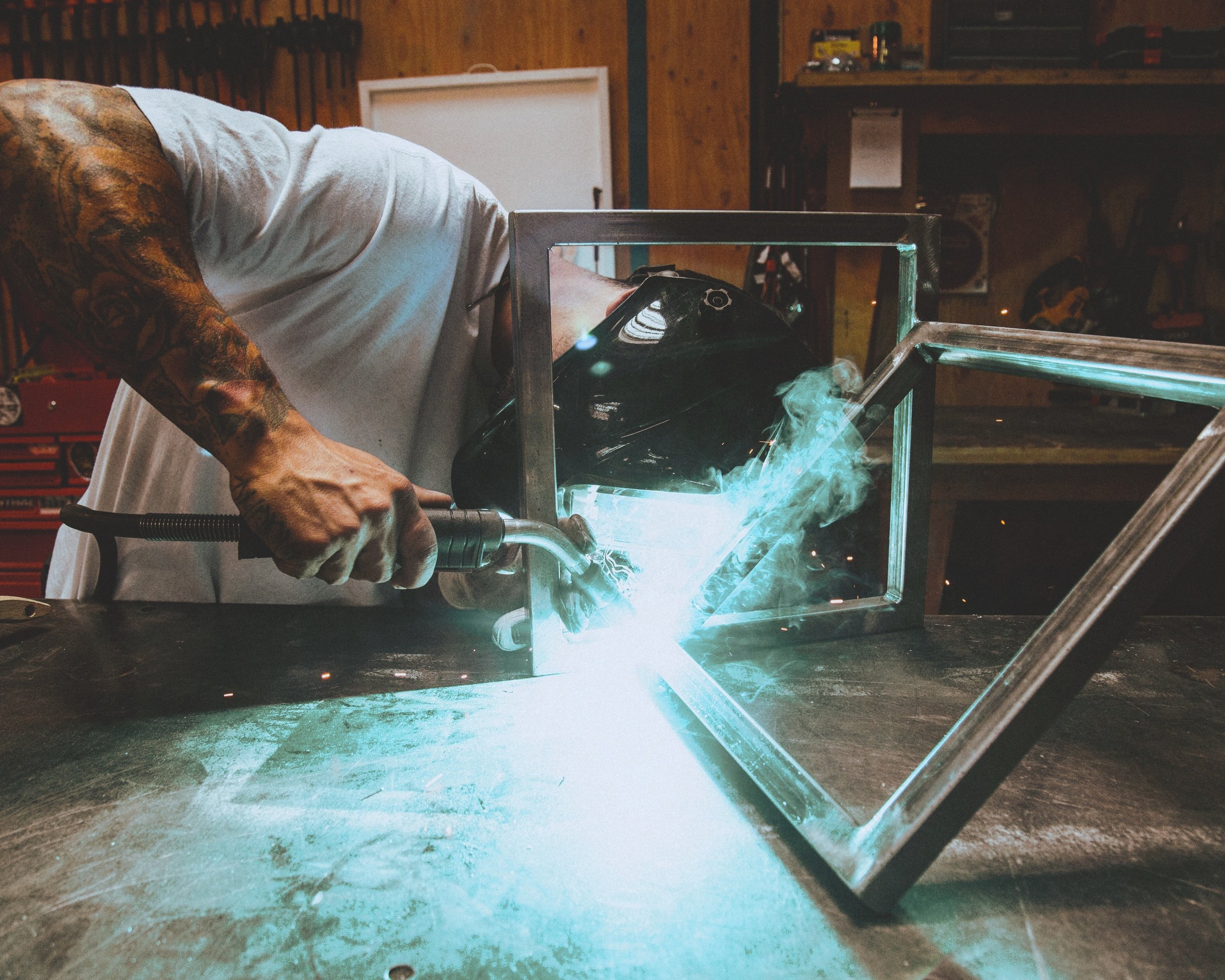 Person welding metal frames in a workshop, wearing a welding helmet, with welding torch in hand, sparks and smoke visible.