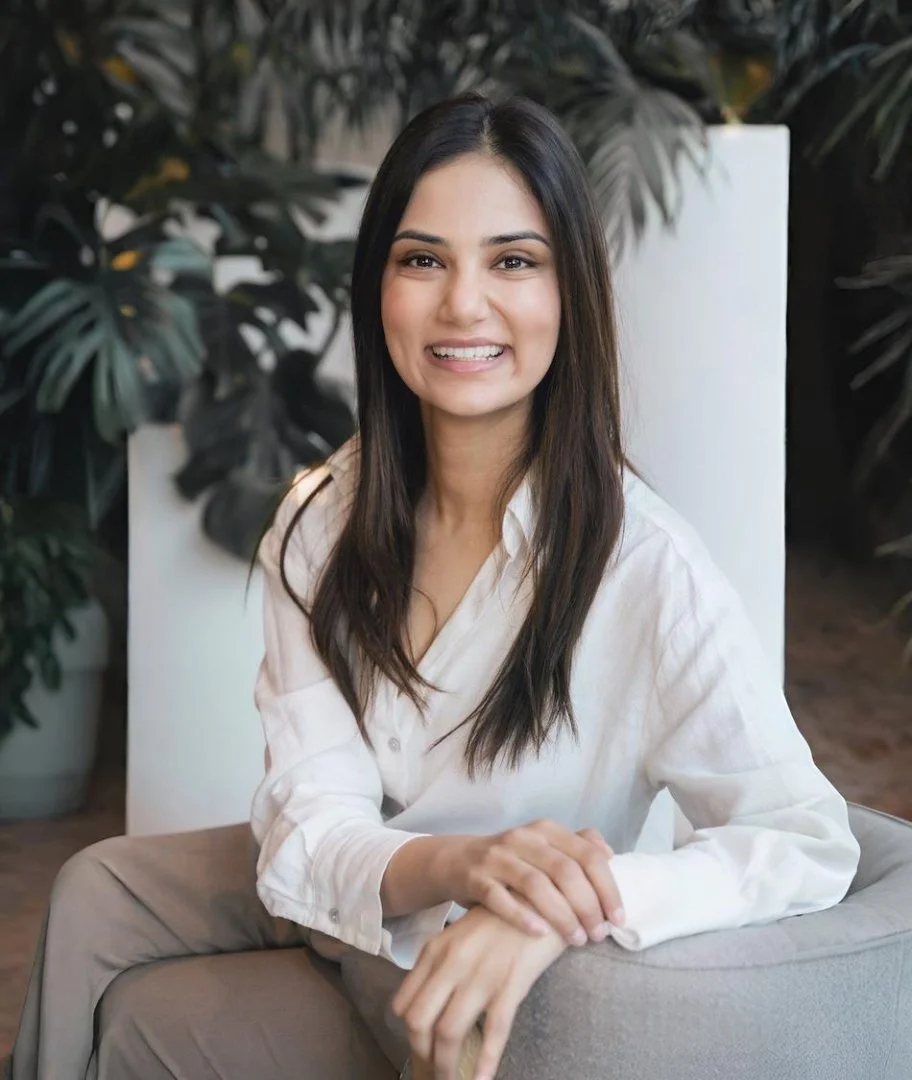 A young woman with long dark hair smiling, sitting on a chair with her hands resting on her lap, in front of leafy green plants.
