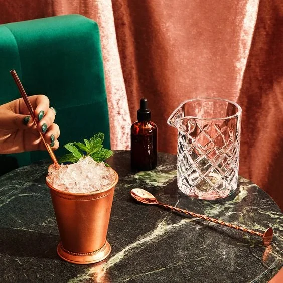 A copper cup filled with a drink and crushed ice, garnished with a mint sprig, on a dark marble table, with a hand holding a copper straw. Nearby are a glass water pitcher, a small amber dropper bottle, and a long copper spoon, against a background o