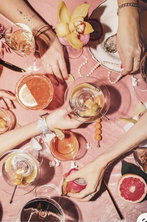 Top-down view of a social gathering with multiple glasses of drinks, some with lemon slices, and hands reaching for a pink grapefruit on a pink fabric surface. There are also pieces of grapefruit and a yellow flower on the table.