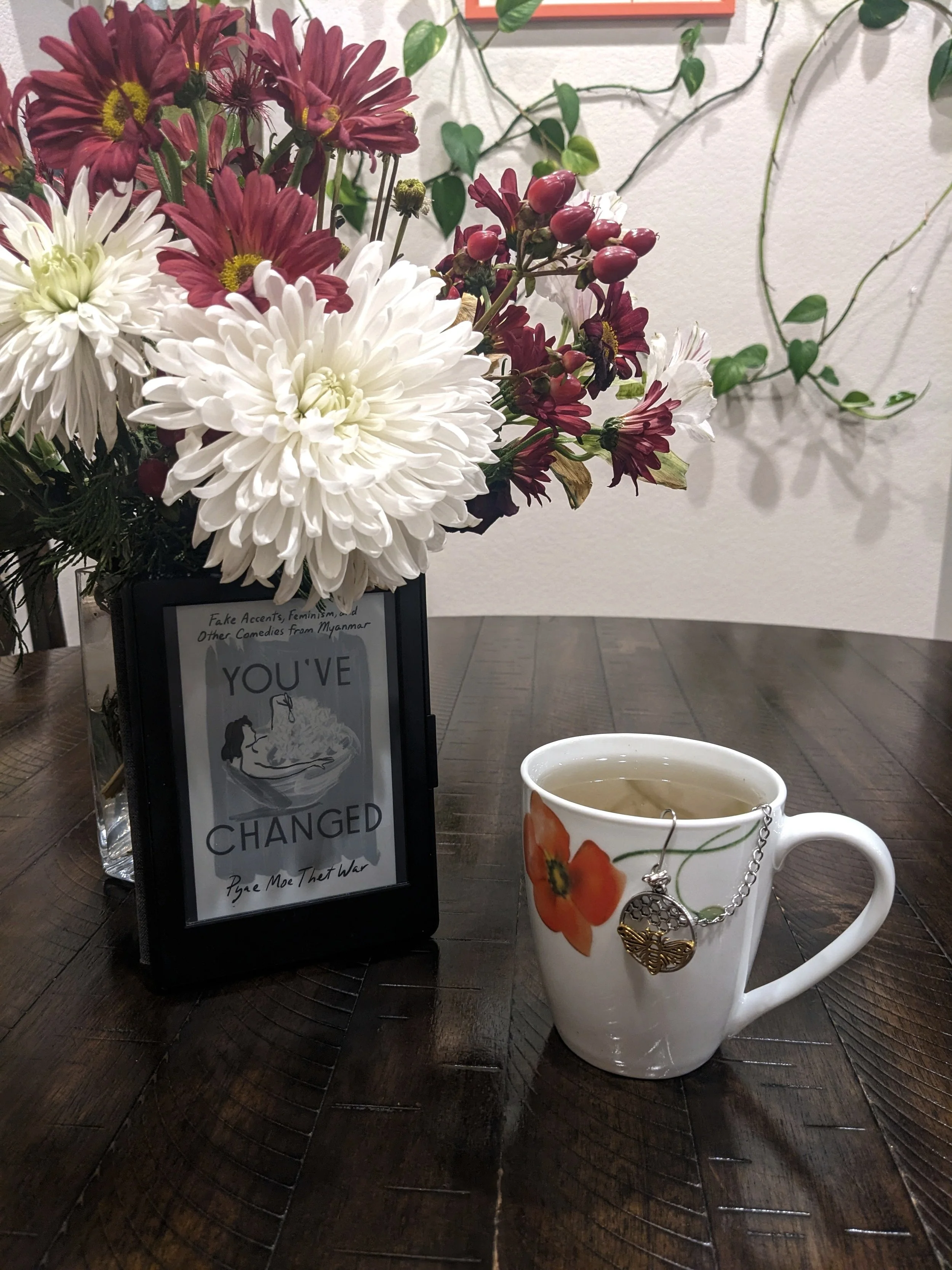 A kindle showing the cover of You've Changed is propped up against a glass vase filled with red and white flowers.  To its right is a white mug printed with an orange poppy filled with tea.  A silver and gold bee charm is hanging from the front of it